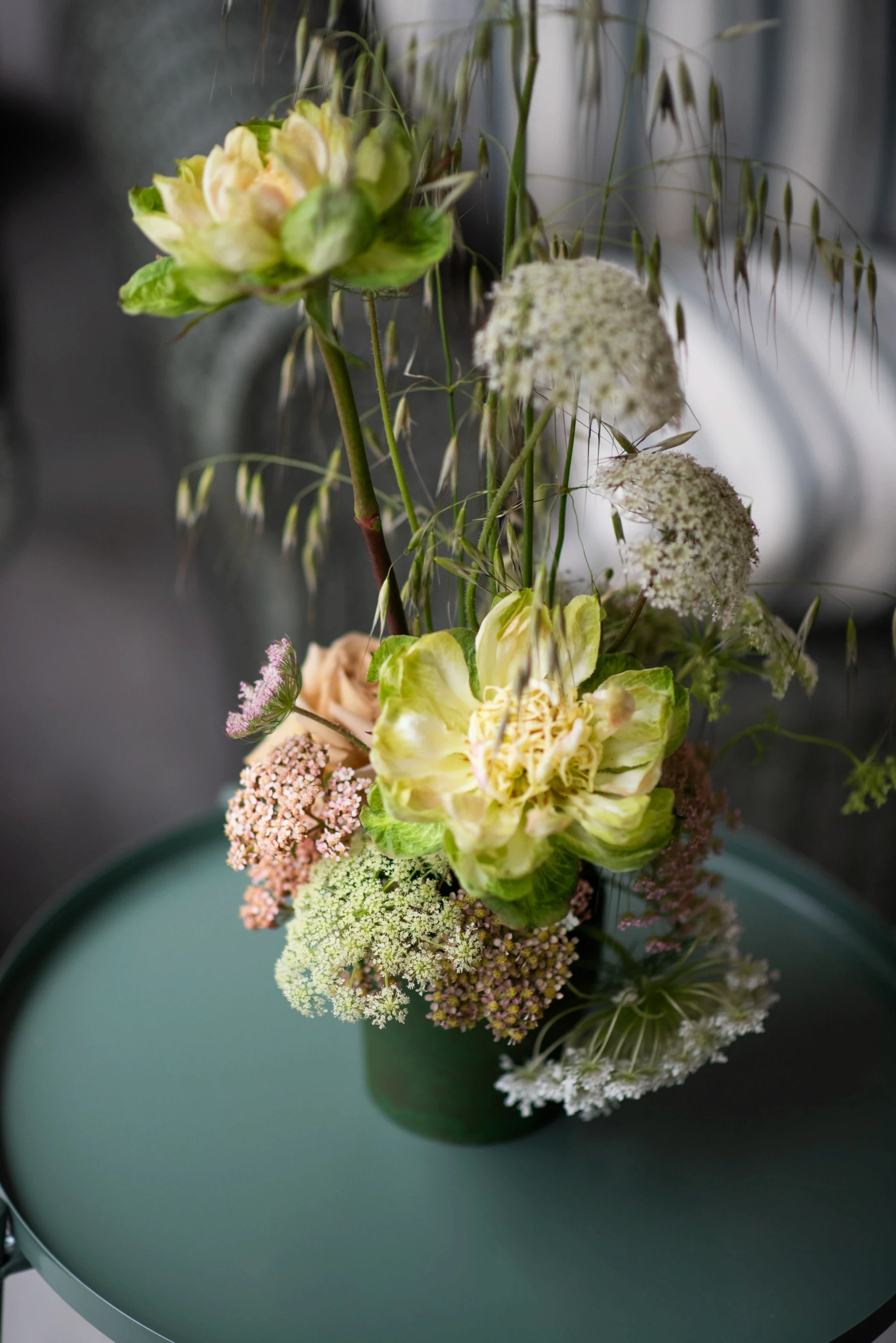 Detalle de florero verde minimalista con flores de encaje blanco y pétalos amarillos sobre mesa circular.