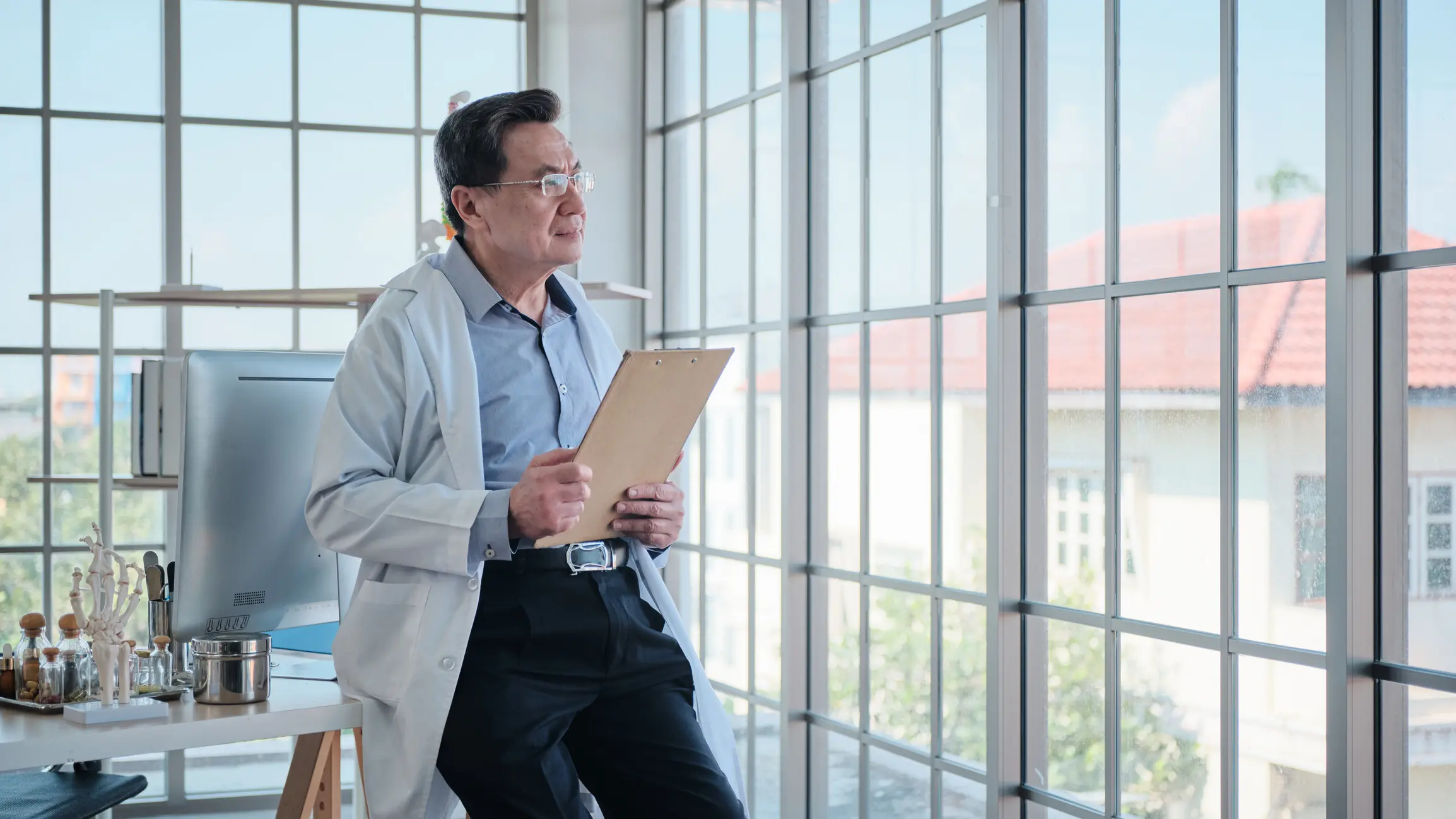 Senior healthcare professional in a white coat standing by a window holding a clipboard, illustrating the reflective space and strategic thinking required for upstream leadership