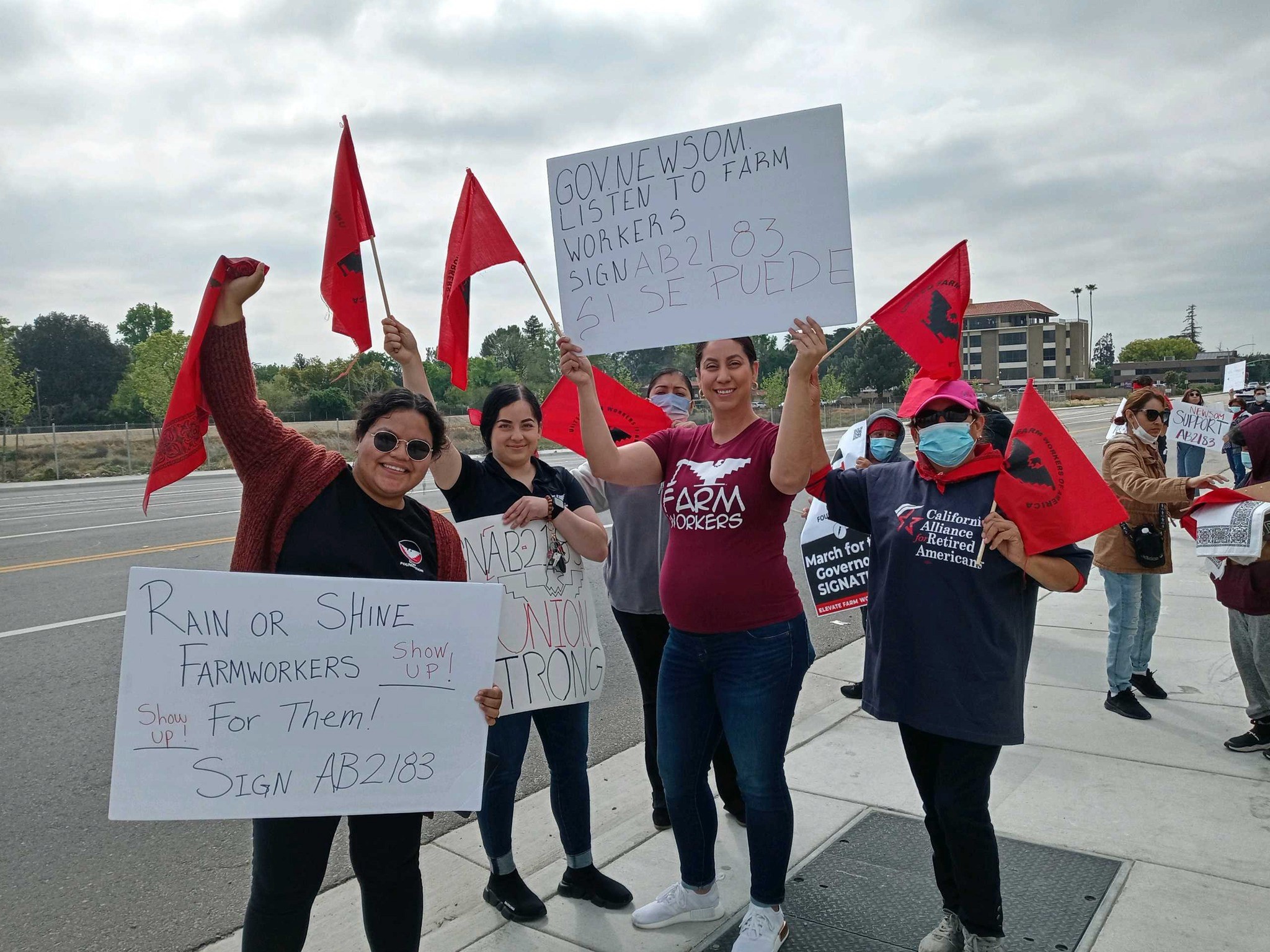 A group of women stand on a sidewalk holding red flags and signs supporting farmworkers' rights and urging the signing of AB 2183. They are smiling and raising fists in solidarity.