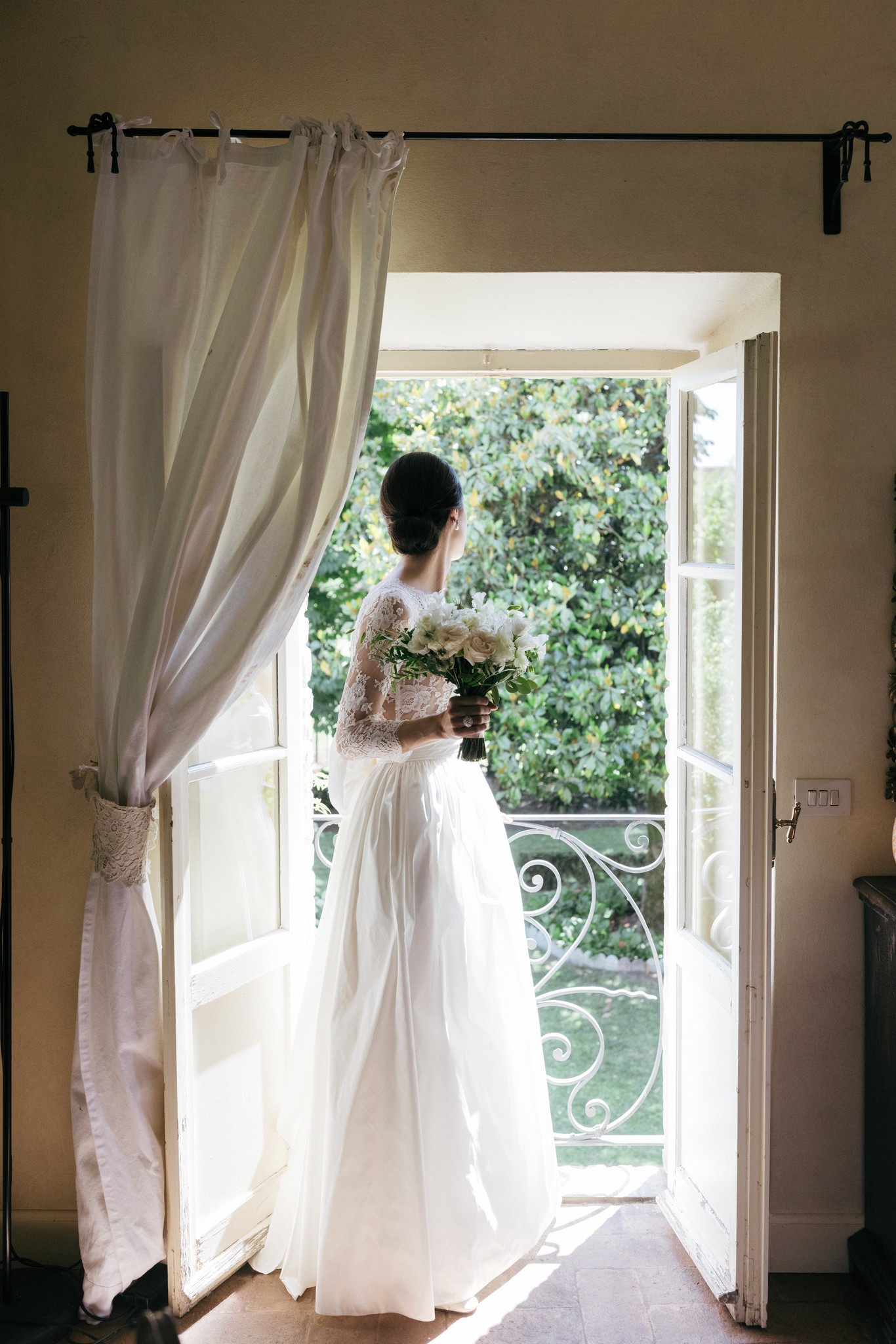 Sposa in abito bianco con bouquet ai preparativi, affacciata alla finestra della villa con tende di lino e vista sul giardino