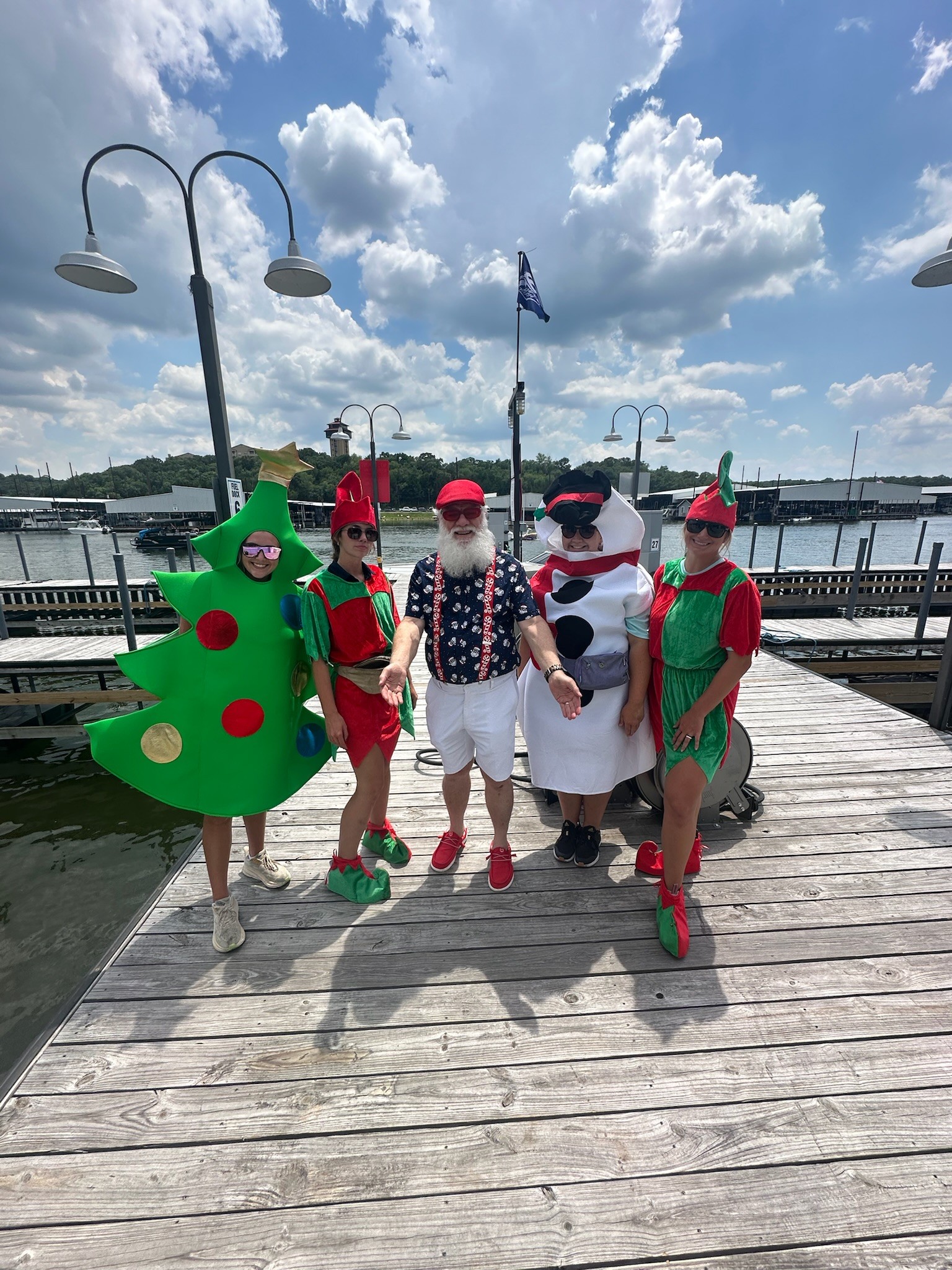 Four people wearing festive costumes, including a Christmas tree, Santa, and a snowman, stand on a wooden dock near a waterfront under a partly cloudy sky.