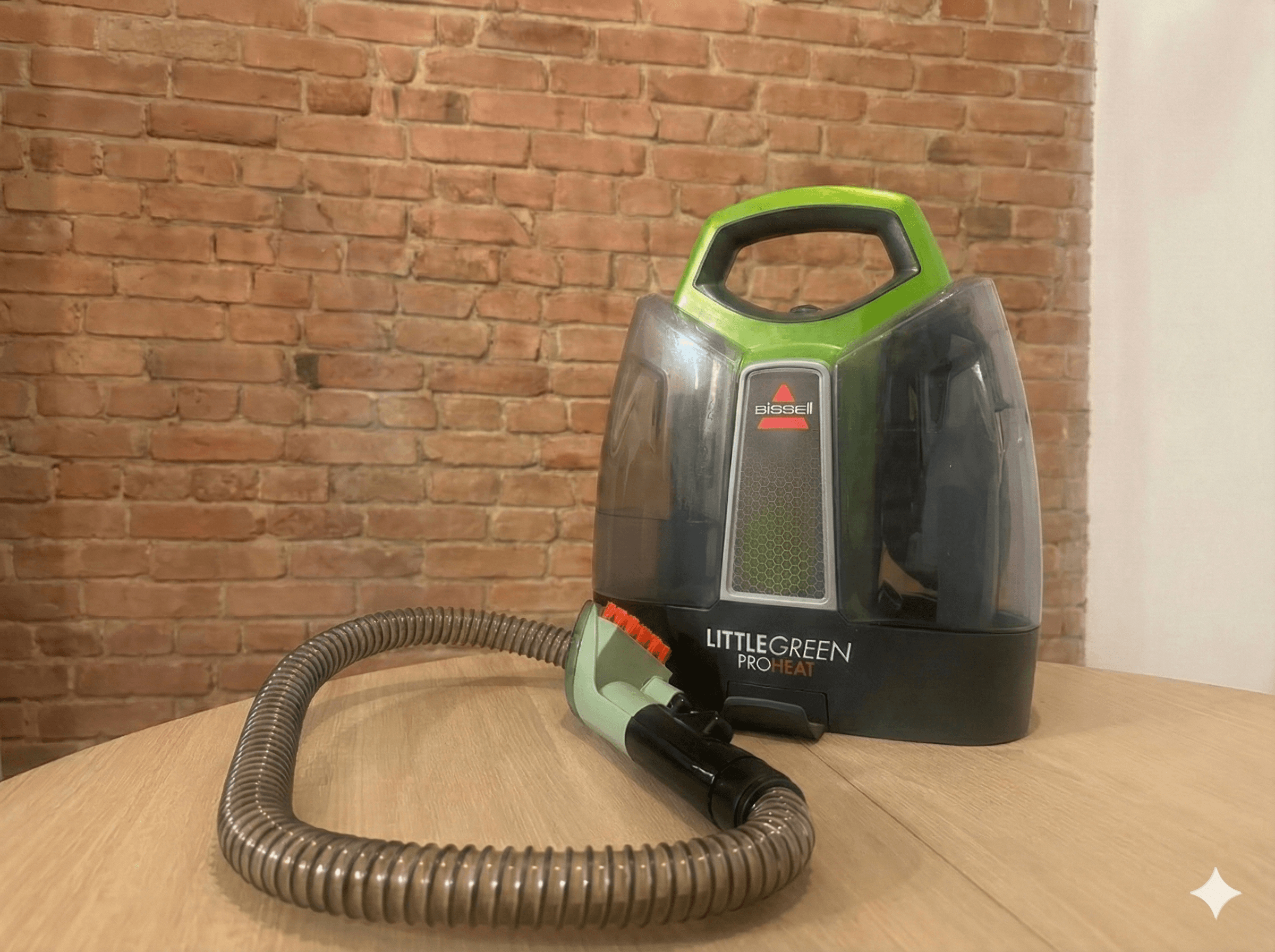 Nettoyeur portatif Bissell Little Green ProHeat posé sur une table en bois, avec un tuyau flexible enroulé, devant un mur de briques apparentes.