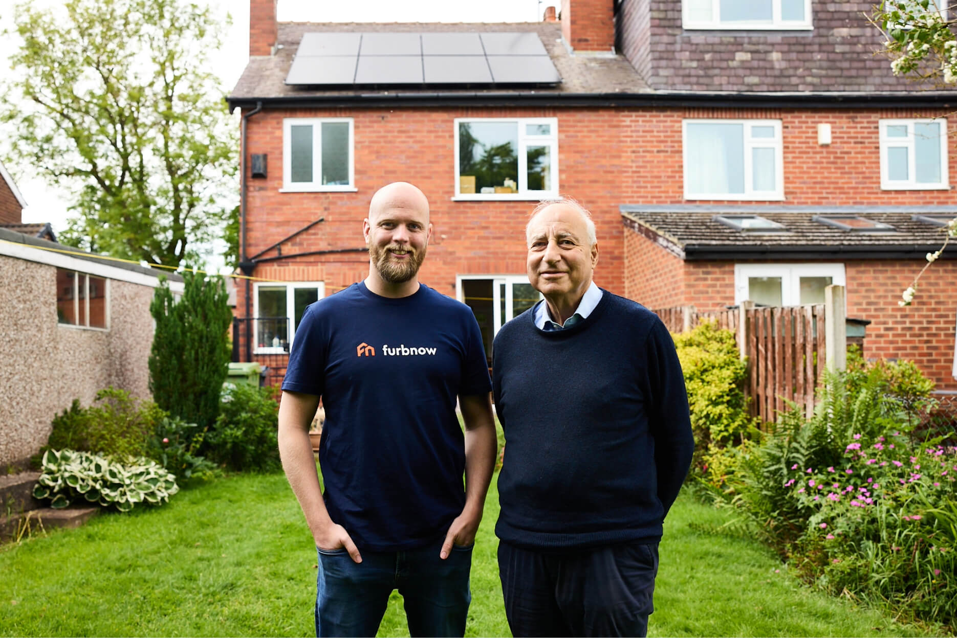 Two people standing and smiling outside a house with solar panels on the roof.