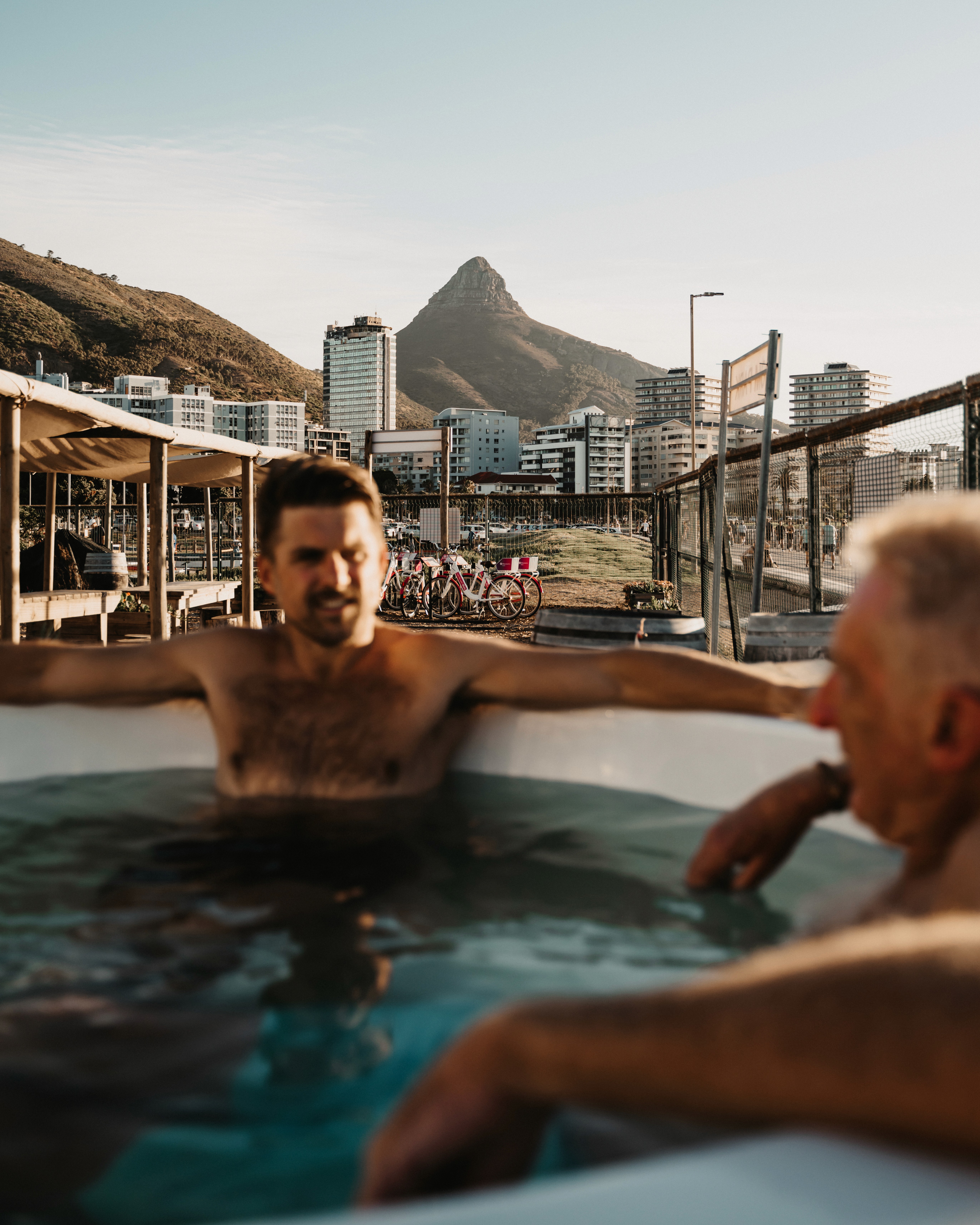Two men relaxing in a cold plunge at SaunaHaüs Cape Town, with Lion’s Head and Sea Point Promenade in the background.