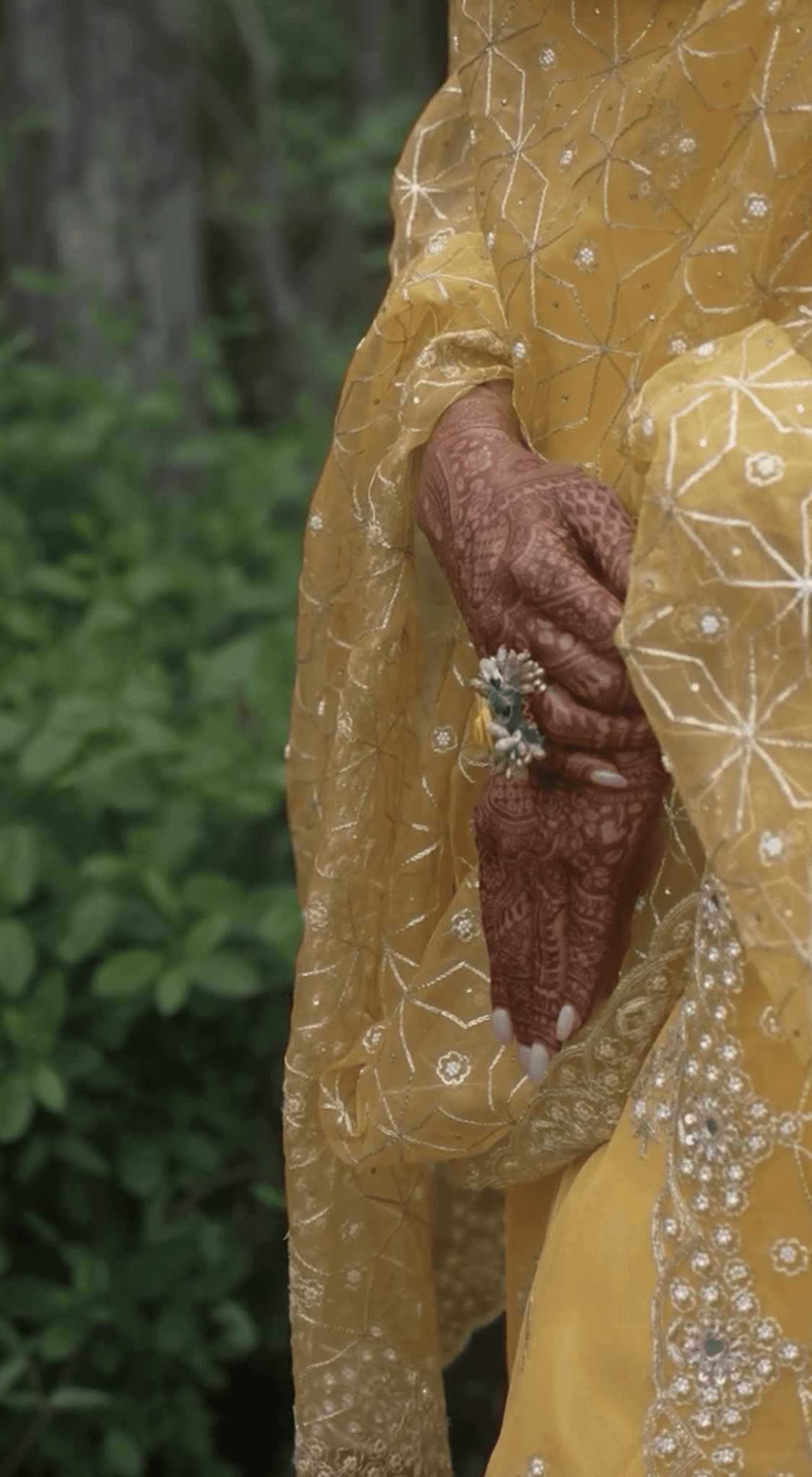 Bride's hand adorned with henna and a floral ring, wearing a yellow and gold dress, captured in an elegant cinematic wedding moment.