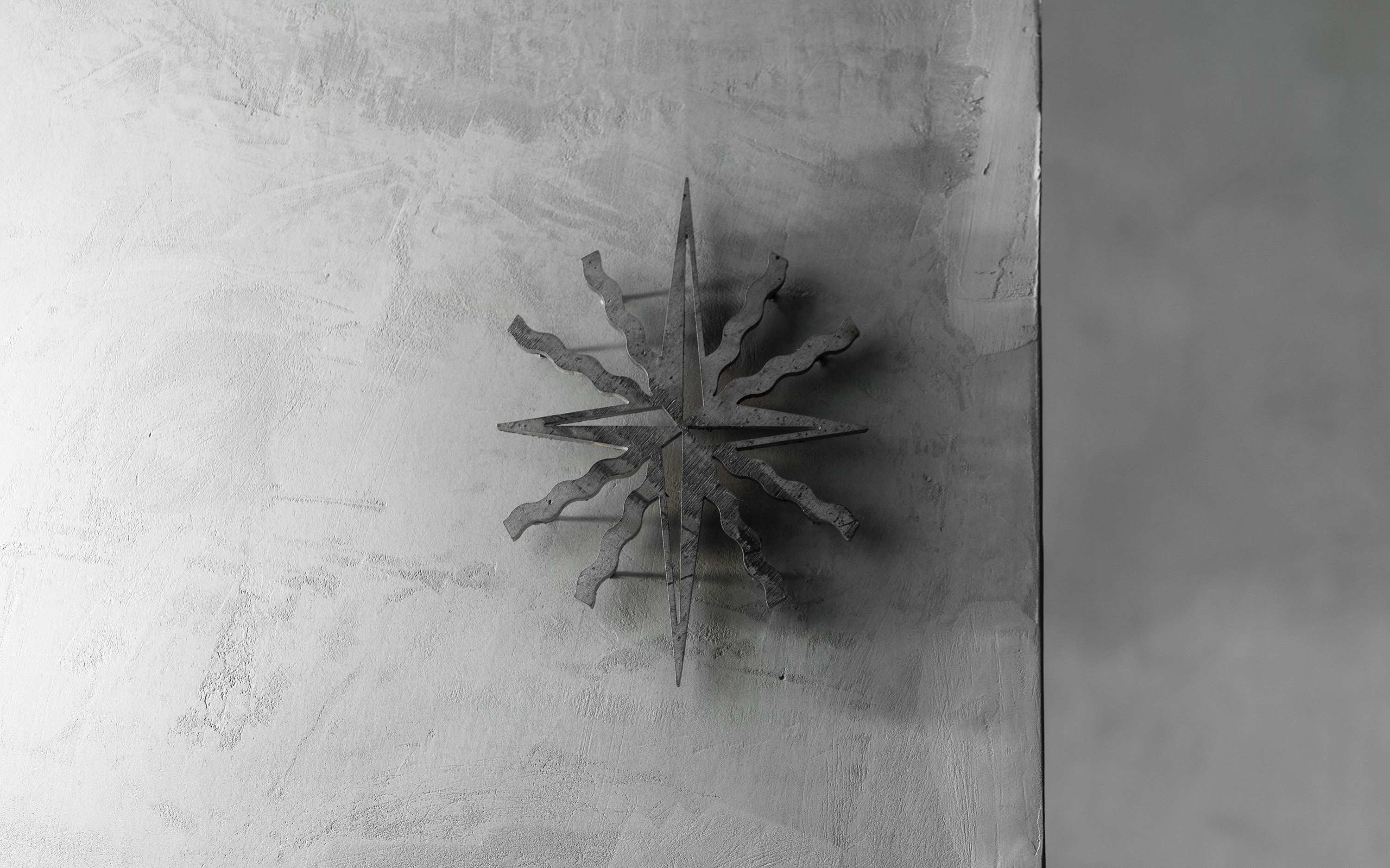 Metallic starburst emblem mounted on matte gray wall with subtle shadow, evoking East by West restaurant's signature motif in industrial style.