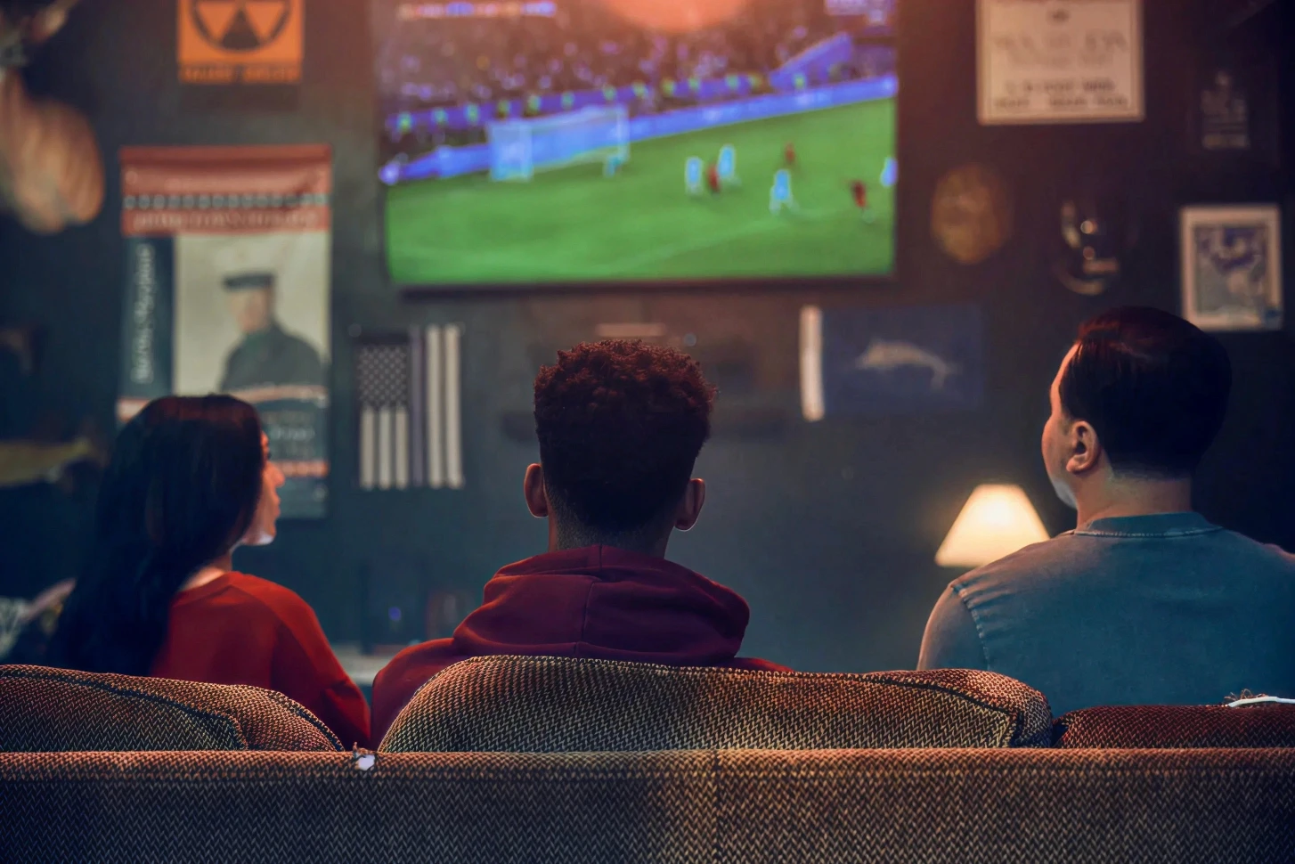 Three friends watching live football match on large wall-mounted TV from behind on sofa in dimly lit sports bar decorated with vintage posters and memorabilia, warm atmospheric lighting