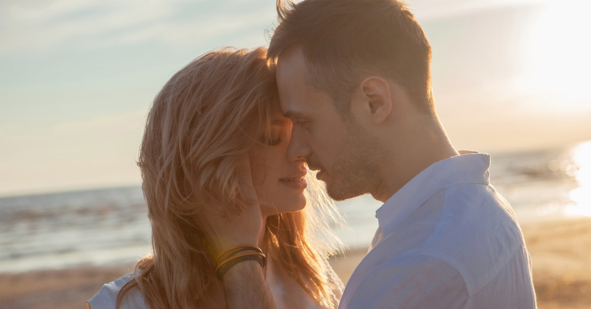 En man och en kvinna är på väg att kyssa varandra, medan mannen har sin hand på kvinnans ansikte. De står på en strand vid solnedgången.