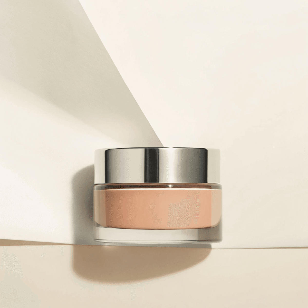 A sleek glass jar with a silver metallic lid, filled with peach-colored cosmetic cream, is elegantly placed on folded, light beige paper, creating a minimalist and luxurious aesthetic.