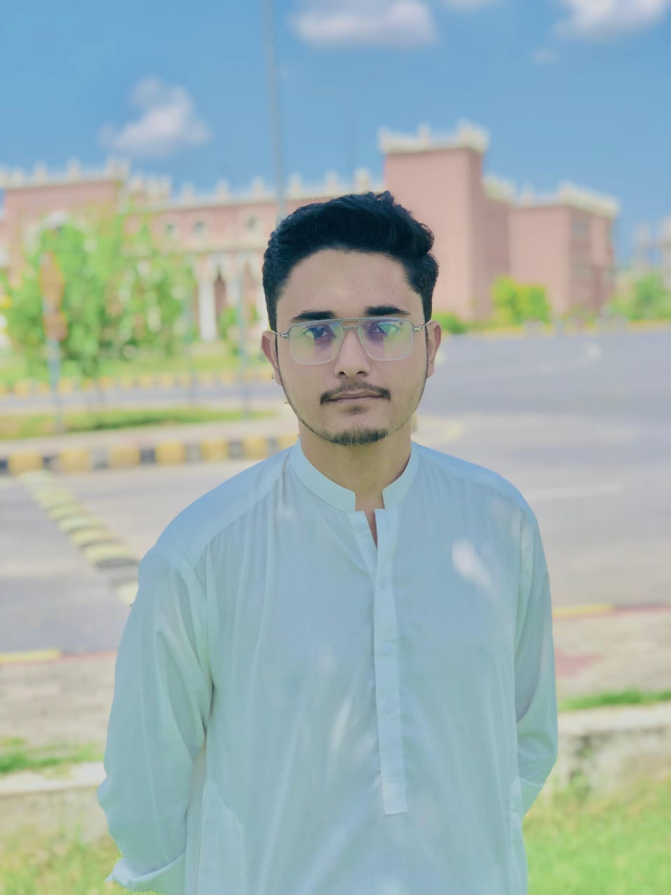 A person wearing a white traditional outfit stands outside on a sunny day, with a blurred historic brick building featuring battlements in the background.