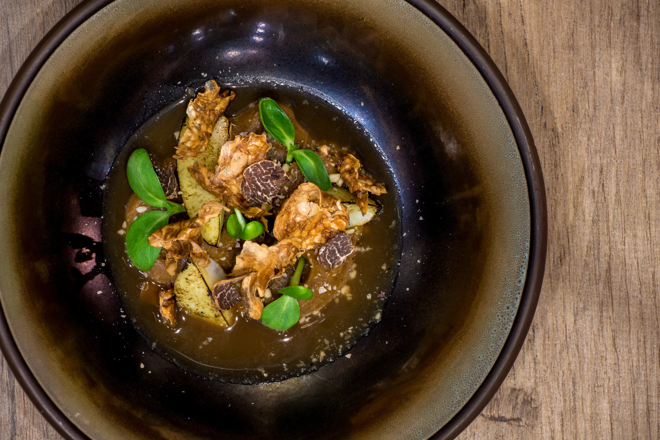 A top-down view of a dark ceramic bowl containing braised meat and green peppers in a savory broth on a wooden table.