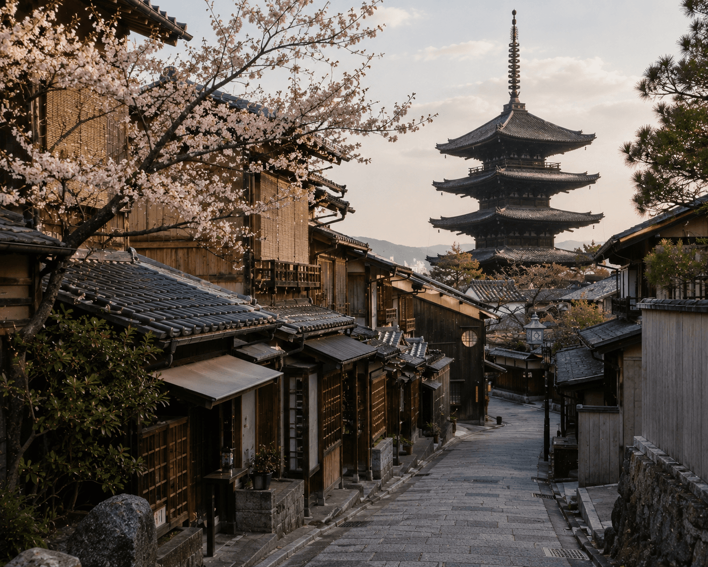 A scenic view looking down a narrow stone-paved street in the historic Higashiyama district of Kyoto, Japan. Traditional dark wooden machiya townhouses line the pathway, with soft pink cherry blossoms blooming from a tree branch in the upper left foreground and the iconic five-story Yasaka Pagoda (Hokan-ji Temple) towering in the background against a soft, sunlit sky.