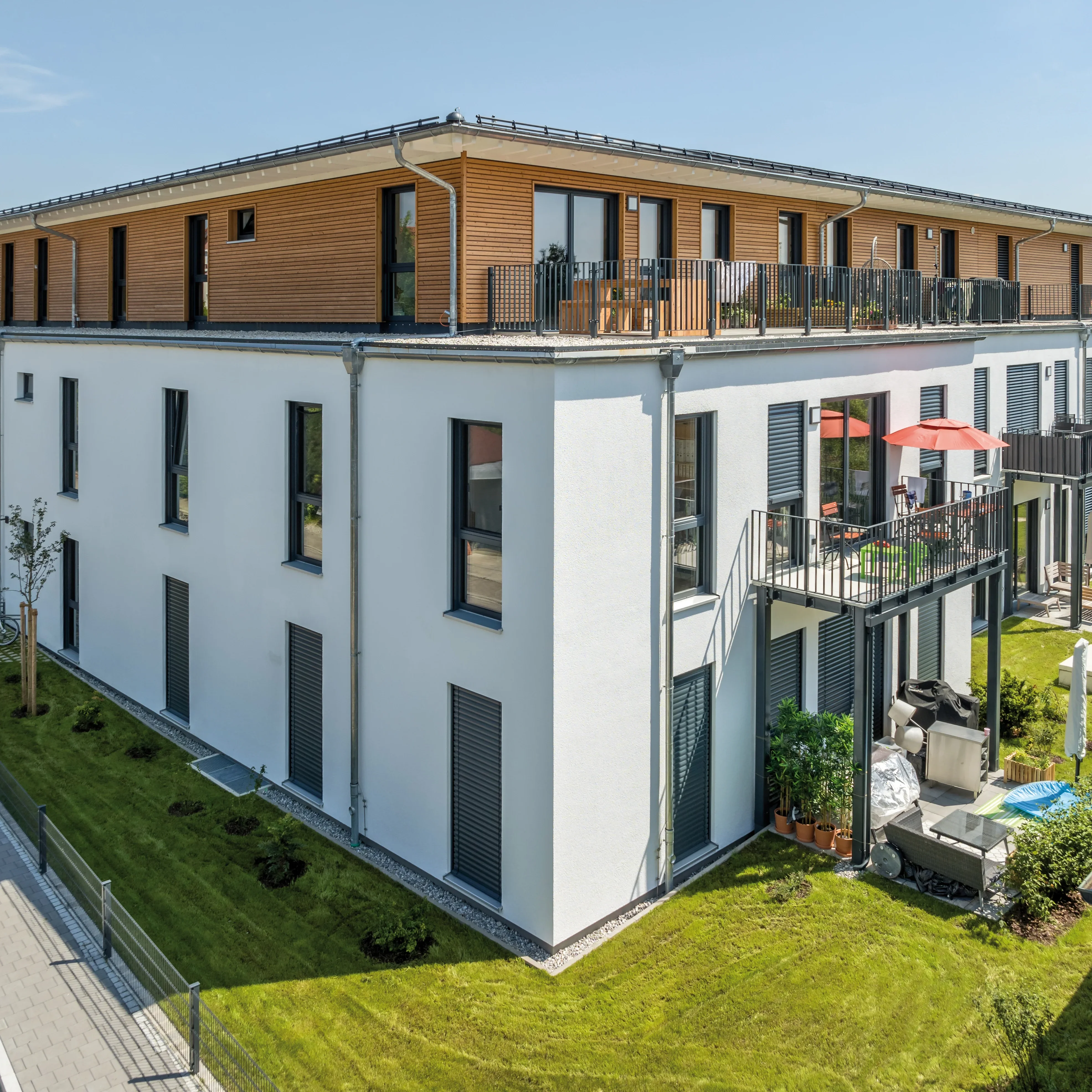 Moderner Mehrfamilienhaus-Eckbau mit weißer Fassade, Holzelementen und Balkonen, umgeben von gepflegten Rasenflächen.