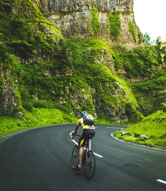 Cyclist wearing a helmet and backpack riding along a winding mountain road surrounded by lush greenery and rocky cliffs.