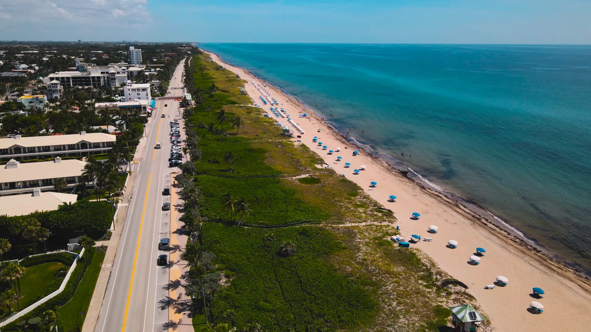 Aerial view of a long sandy beach beside the ocean, with a coastal road and green dunes running parallel to the shoreline.