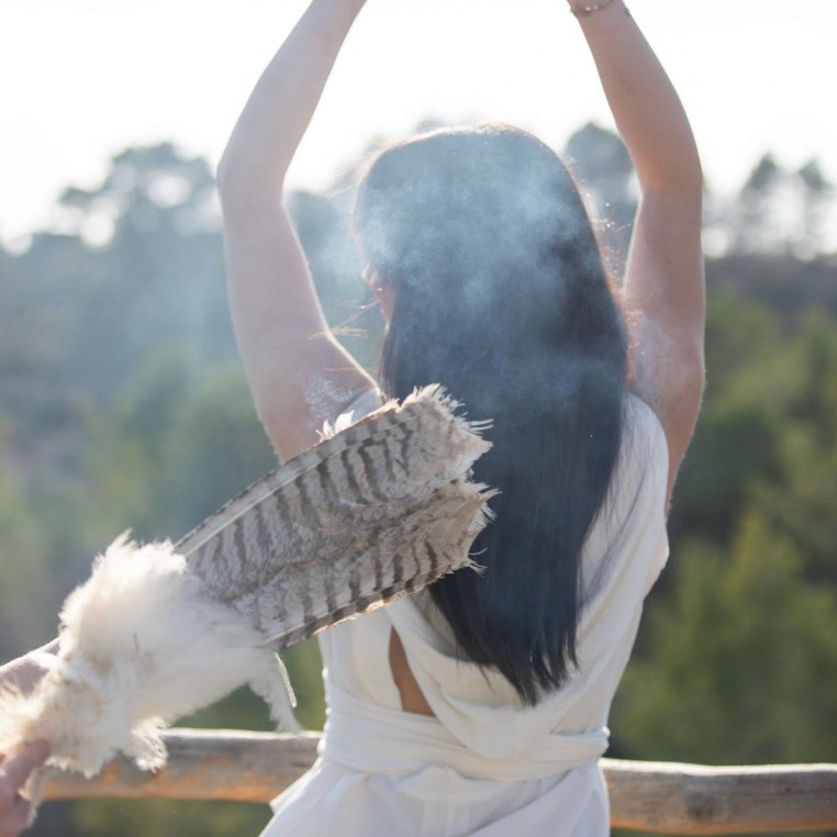 Sunoom Resort | A person with long dark hair stands outdoors, arms raised, holding feathers, with smoke gently surrounding them.