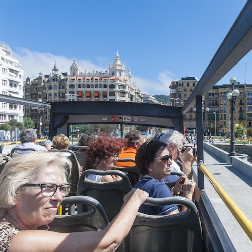 Passengers seated on the upper deck of an open-top bus, viewing a riverside city scene with buildings and greenery.