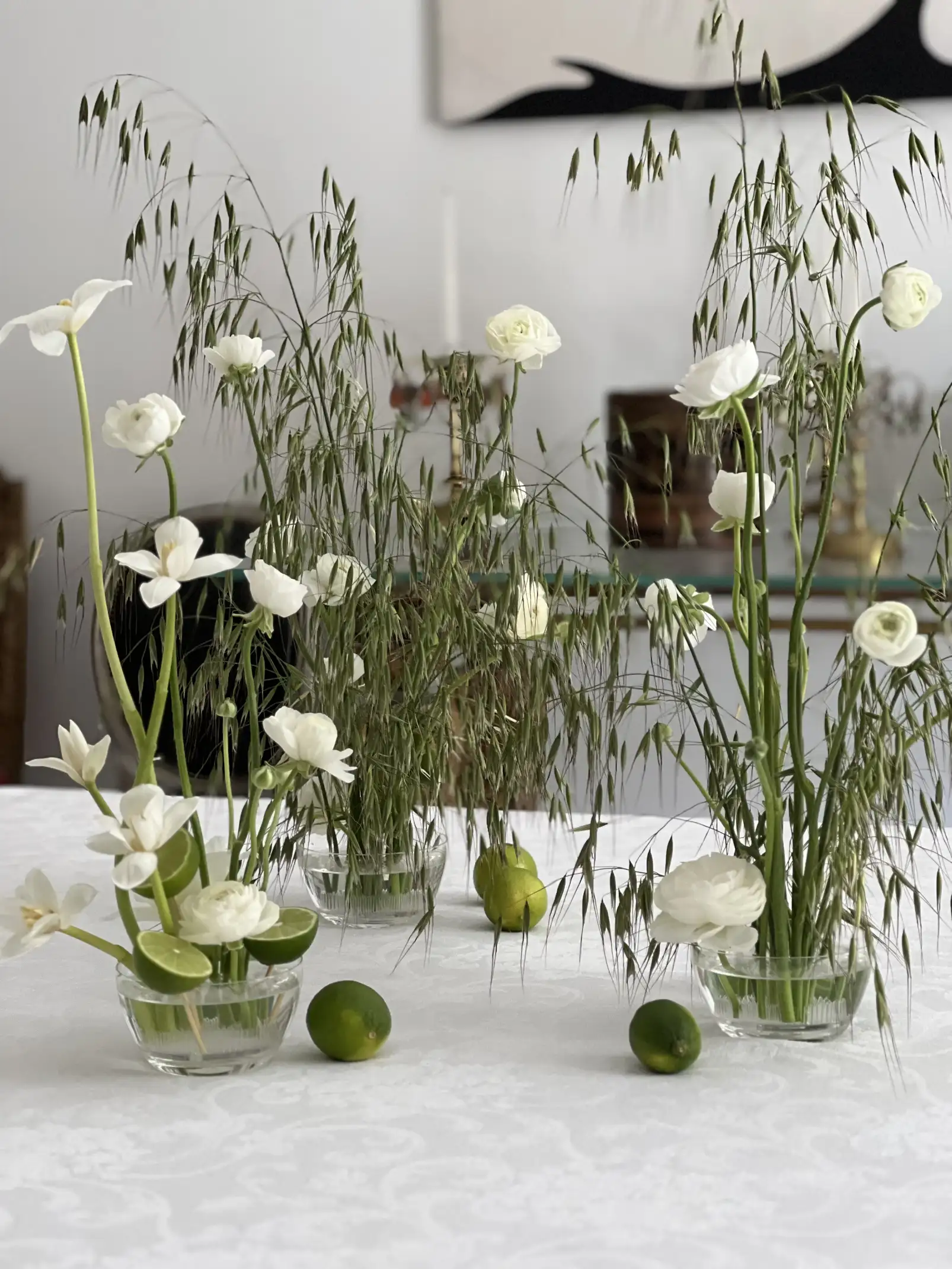 Composición minimalista de arreglos florales con flores blancas y espigas verdes en pequeños floreros de vidrio, intercalados con limas frescas sobre un mantel blanco, creando una escena natural y equilibrada con estética contemporánea.
