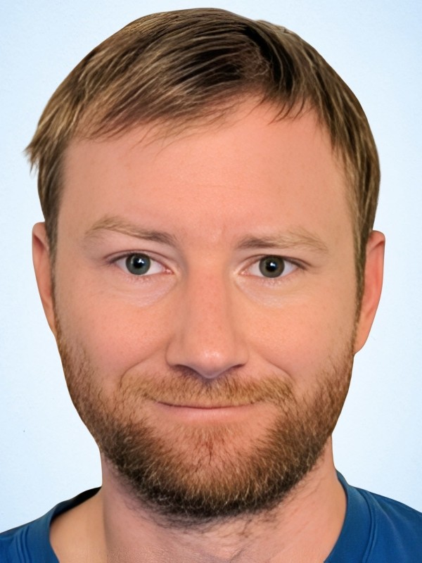 Smiling man with a beard in a dark suit and tie, standing against a gray background. He appears confident and professional.