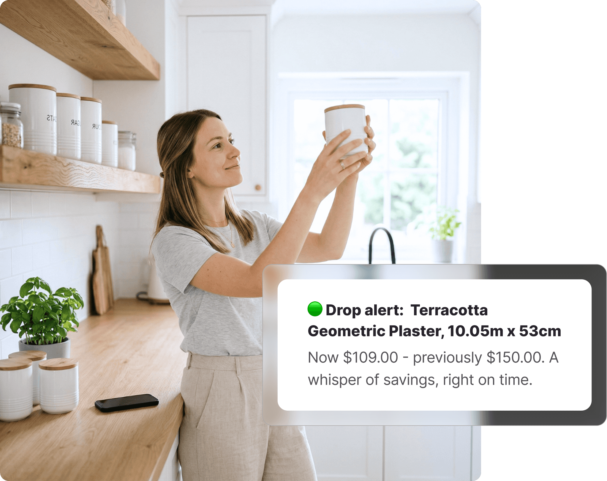 Woman in a bright kitchen holding up a white ceramic canister and examining it, with matching Myer-style kitchen accessories on shelves and a phone on the counter, receiving a price drop alert on Whisprice.