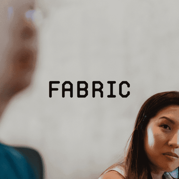 The image presents a close-up view of a woman's face alongside a text that reads "FABRIC." The woman is positioned on the right side of the image, with only a portion of her face visible. She has long brown hair and appears to be wearing a white top. The text "FABRIC" is prominently displayed in black at the center of the image.  To the left of the woman is a partially visible figure who seems to be wearing a blue jacket. However, the detail faces away from the camera, making it difficult to distinguish any specific features.  The background of the image is blurry, with white curtains and beige tones resonating throughout. The overall effect of the image is soft and delicate, with a subtle play of textures and colors that draw the viewer's attention to the central figure of the woman.