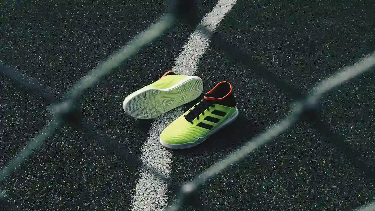 A pair of neon green indoor football shoes lying on a black artificial turf pitch, photographed through a chain-link fence – symbolising the fast-paced, urban style of the Baller League UK.