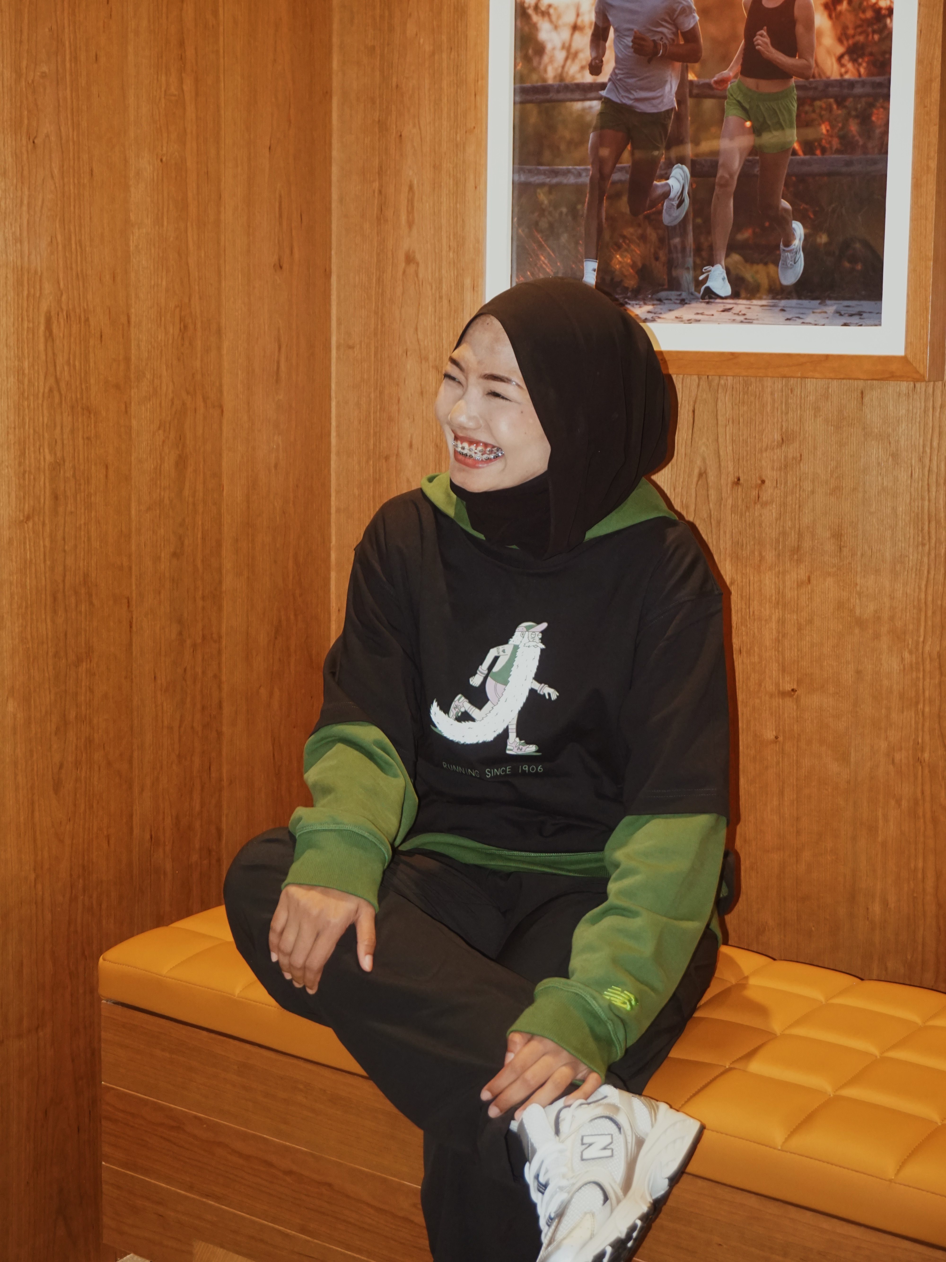 Kak Fit wearing a New Balance black and green athletic outfit sits on a bench in a cozy space, with a motivational running poster in the background.