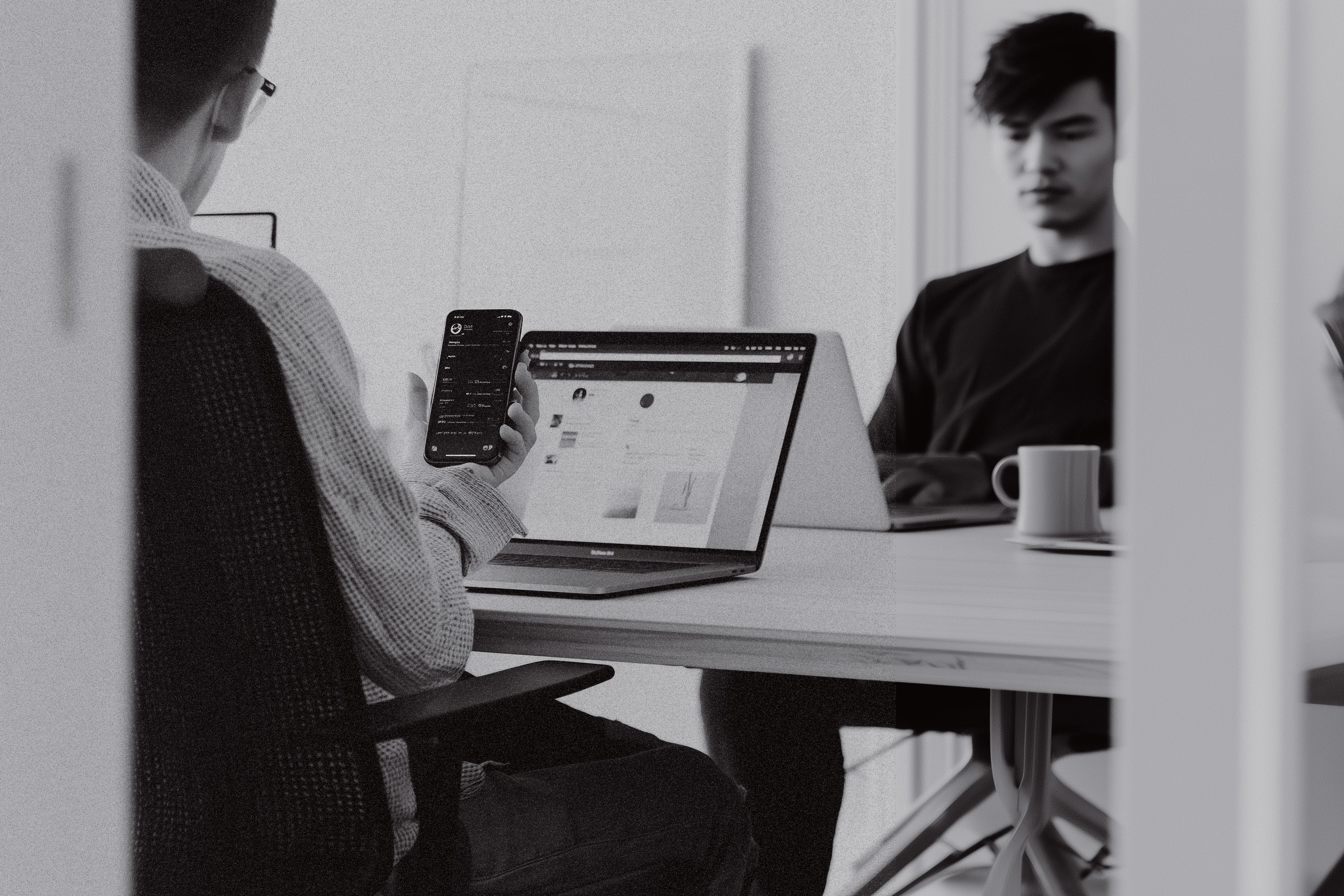 Black-and-white photo of two people working at a table with laptops, one holding a smartphone, with a coffee cup on the desk