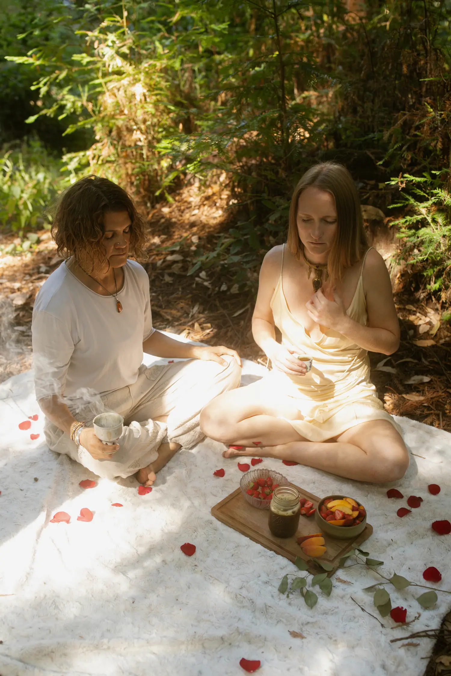 Two women in somatic ceremony seated on a white blanket with rose petals and offerings