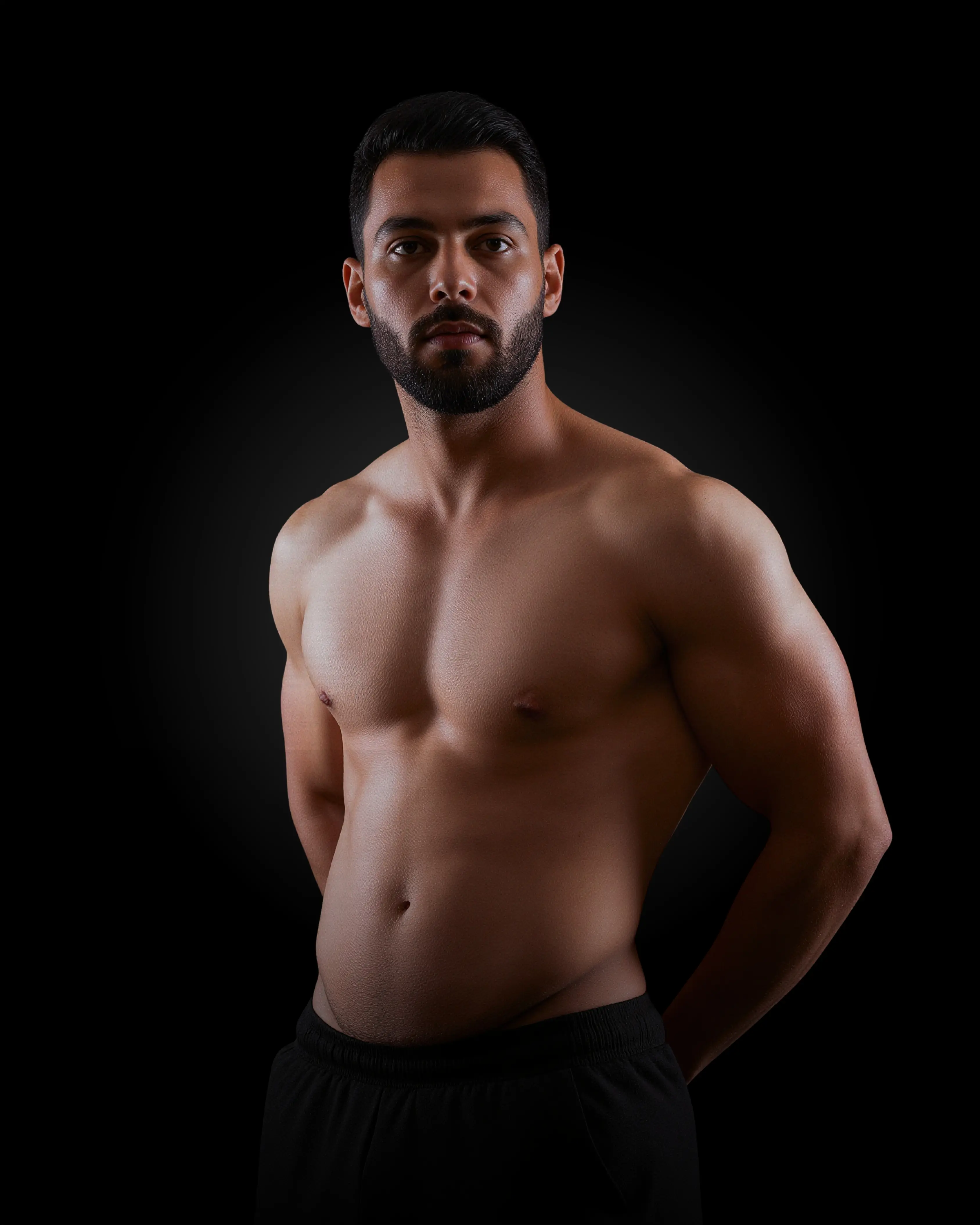 A muscular man with a beard posing shirtless, looking directly at the camera with his hands behind his back.