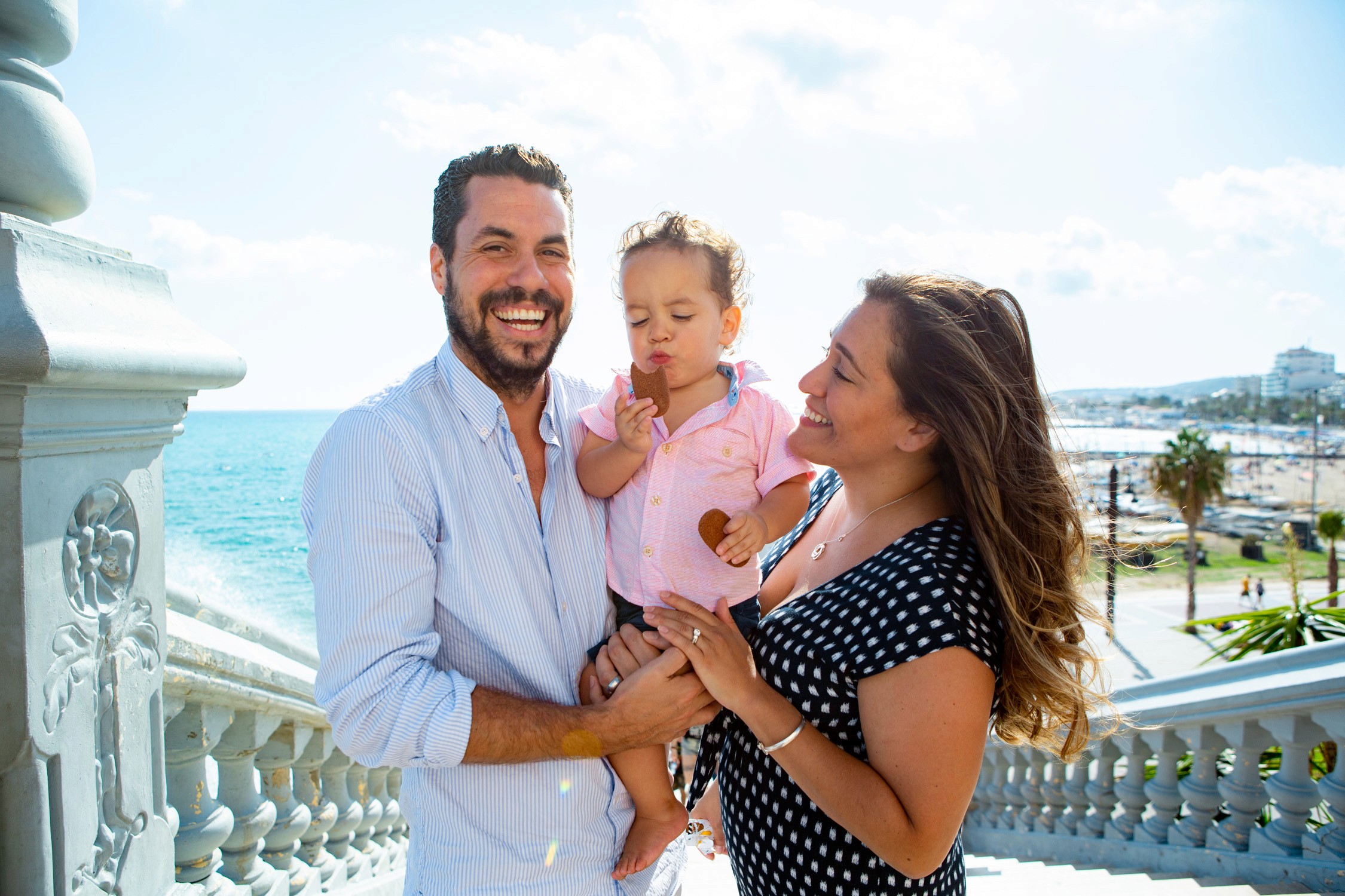 Sunny family enjoys a bright day overlooking the blue ocean from a white balcony railing.