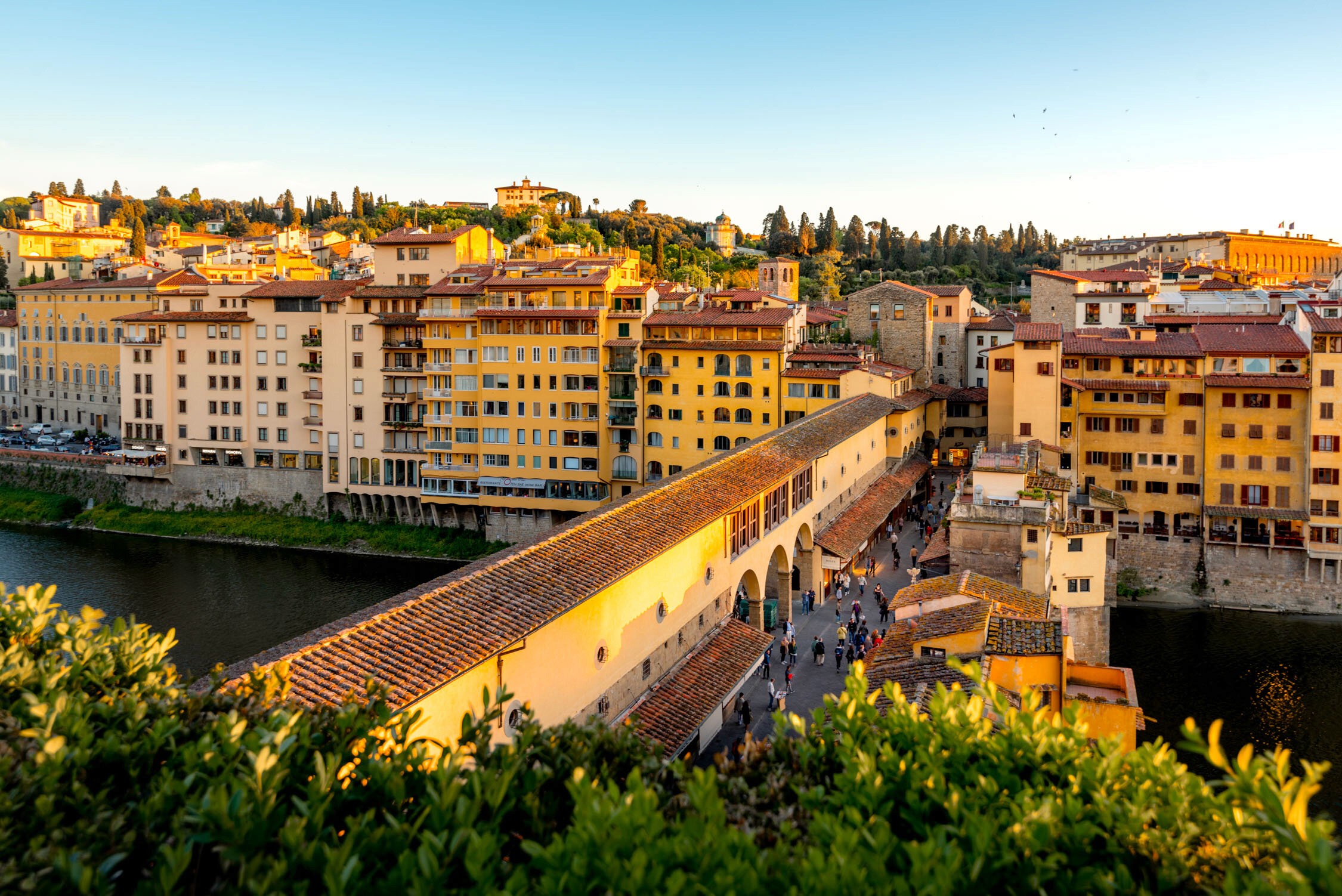 A wide view of a riverside promenade lined with yellow and orange buildings under a clear blue sky at sunset.