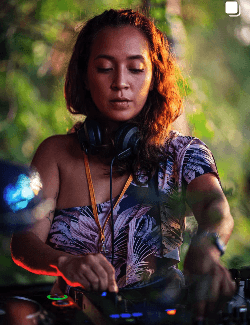 Smiling woman in beige yoga outfit sitting in natural light with green plants in the background.