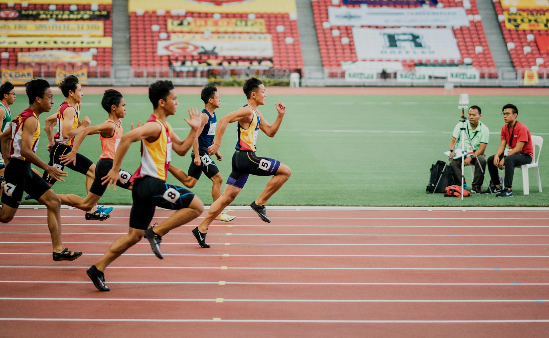 Several male athletes sprint down a track lane during a competitive race with stadium seating in the background.