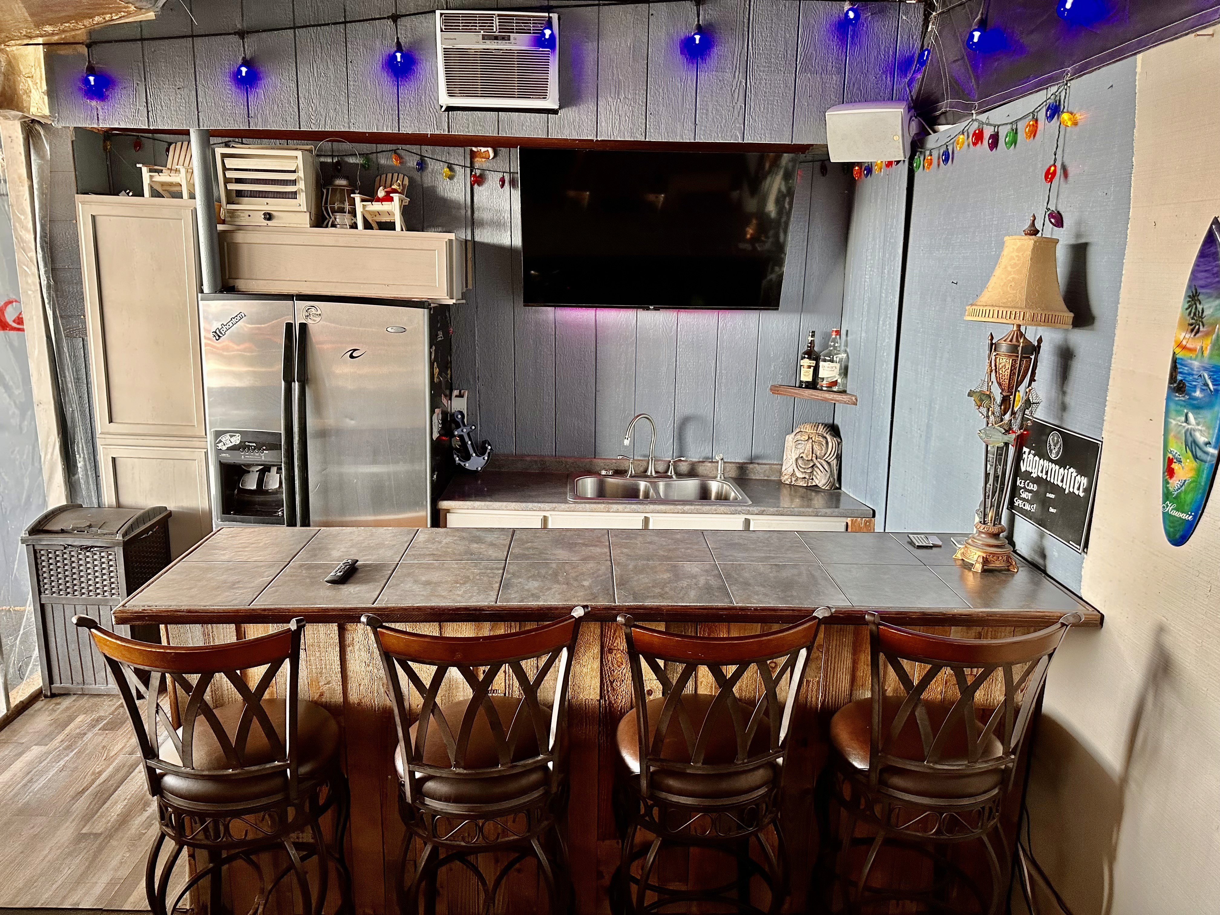 A cozy home bar with wooden chairs lined up at a tile countertop, a mounted flat-screen TV above a stainless steel refrigerator, colorful string lights, and rustic decor accents.