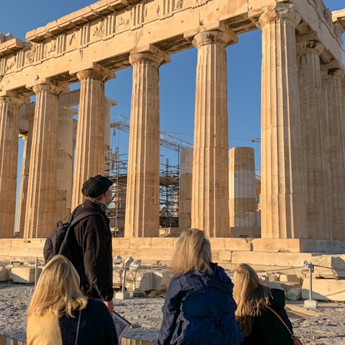 四个人在晴朗的蓝天下观看帕特农神庙，这座古希腊神庙拥有高耸的立柱。内部可见脚手架。