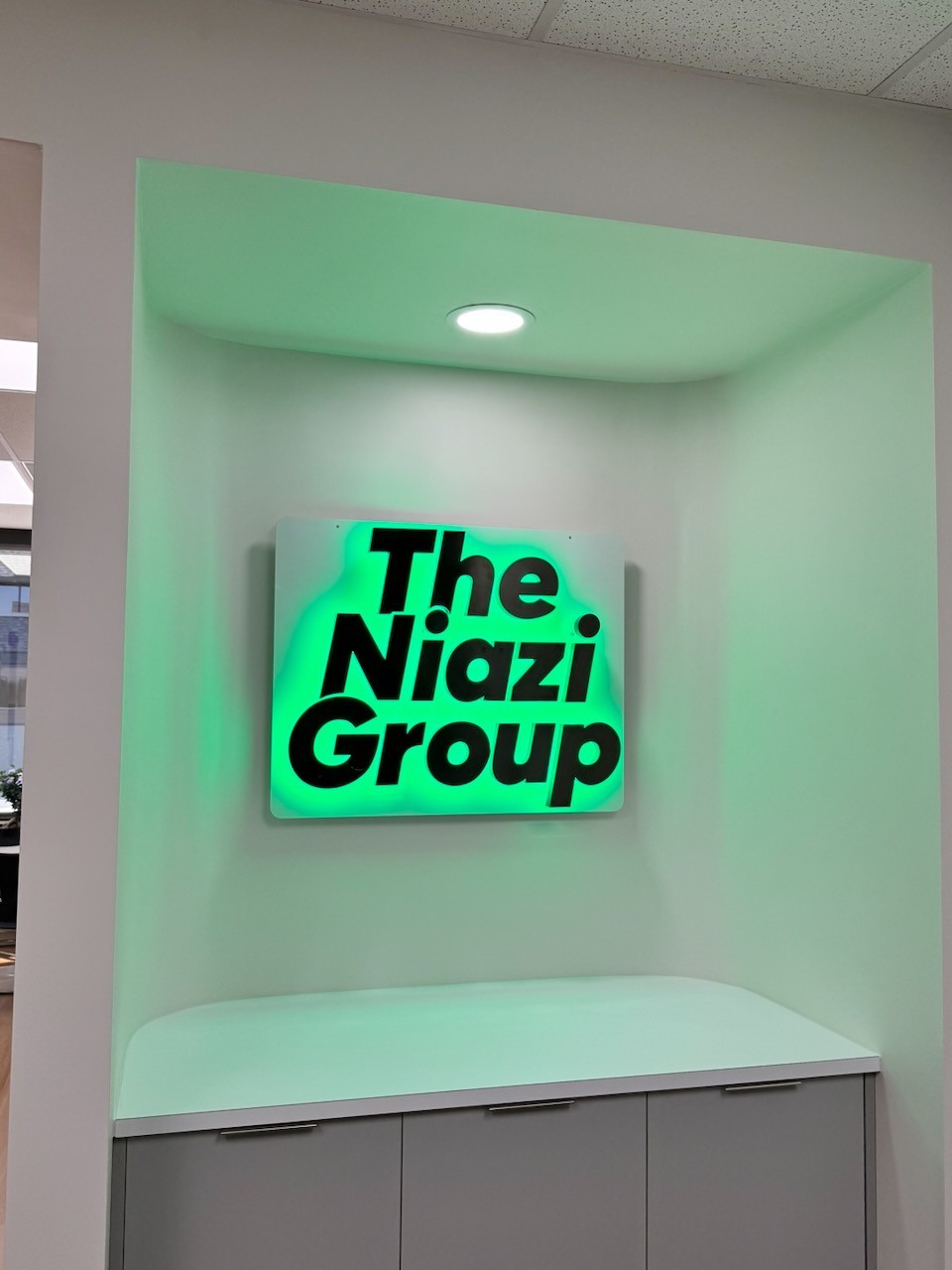 A custom interior lobby sign for The Niazi Group featuring black dimensional lettering on a backlit frosted acrylic panel with vibrant green LED lighting, mounted in a recessed wall alcove.