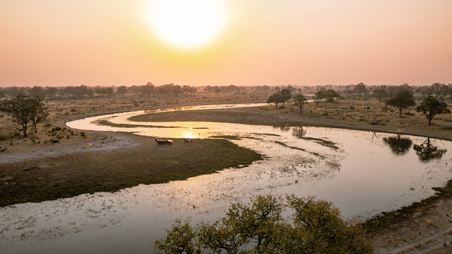 Kujwana Delta Ride, Okavango Delta, Botswana. Drönarvy över fyra personer på game drive som blickar ut över solnedgången, med en safarijeep bredvid sig och en av deltats breda kanaler i förgrunden