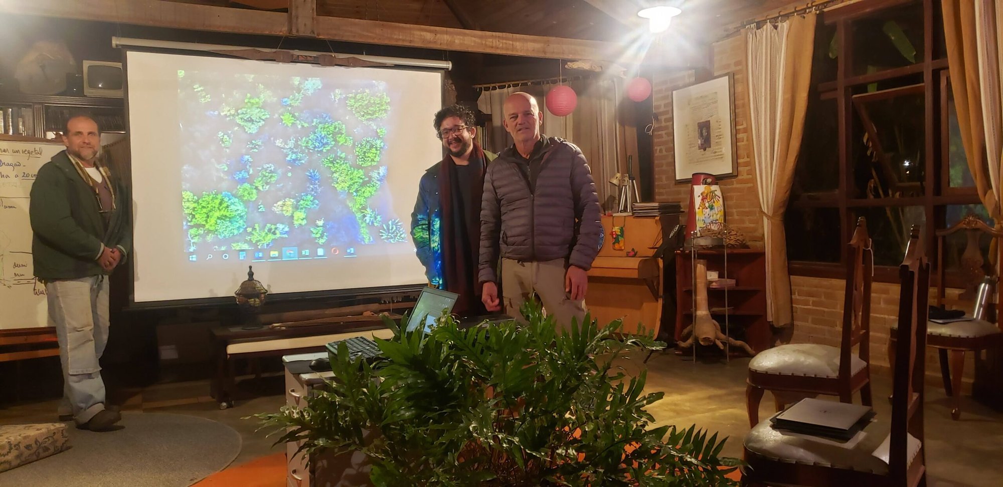 Três homens diante de telão com mapa de satélite em apresentação noturna