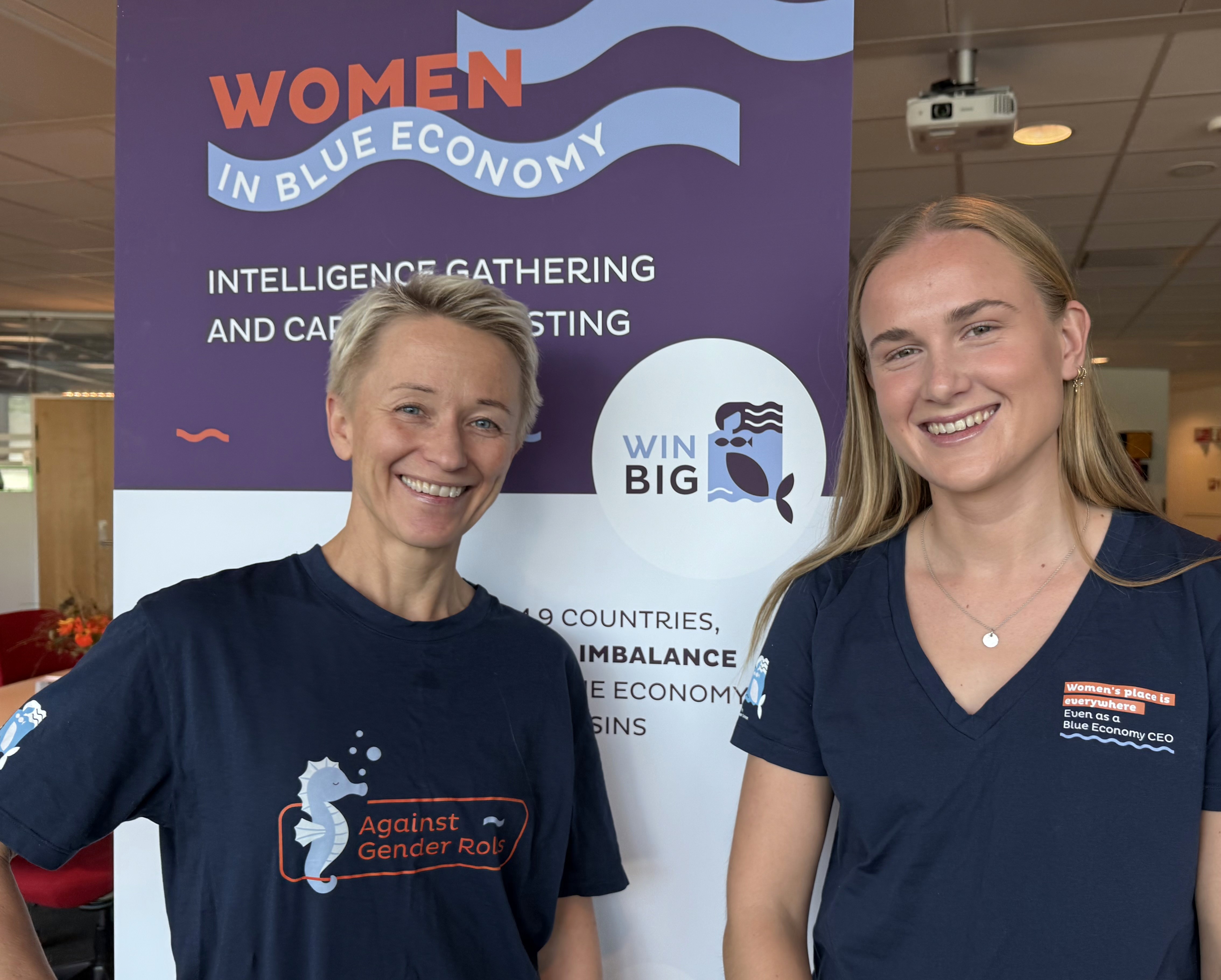 Two women stand smiling in front of a colorful poster. They wear matching t-shirts and appear friendly.