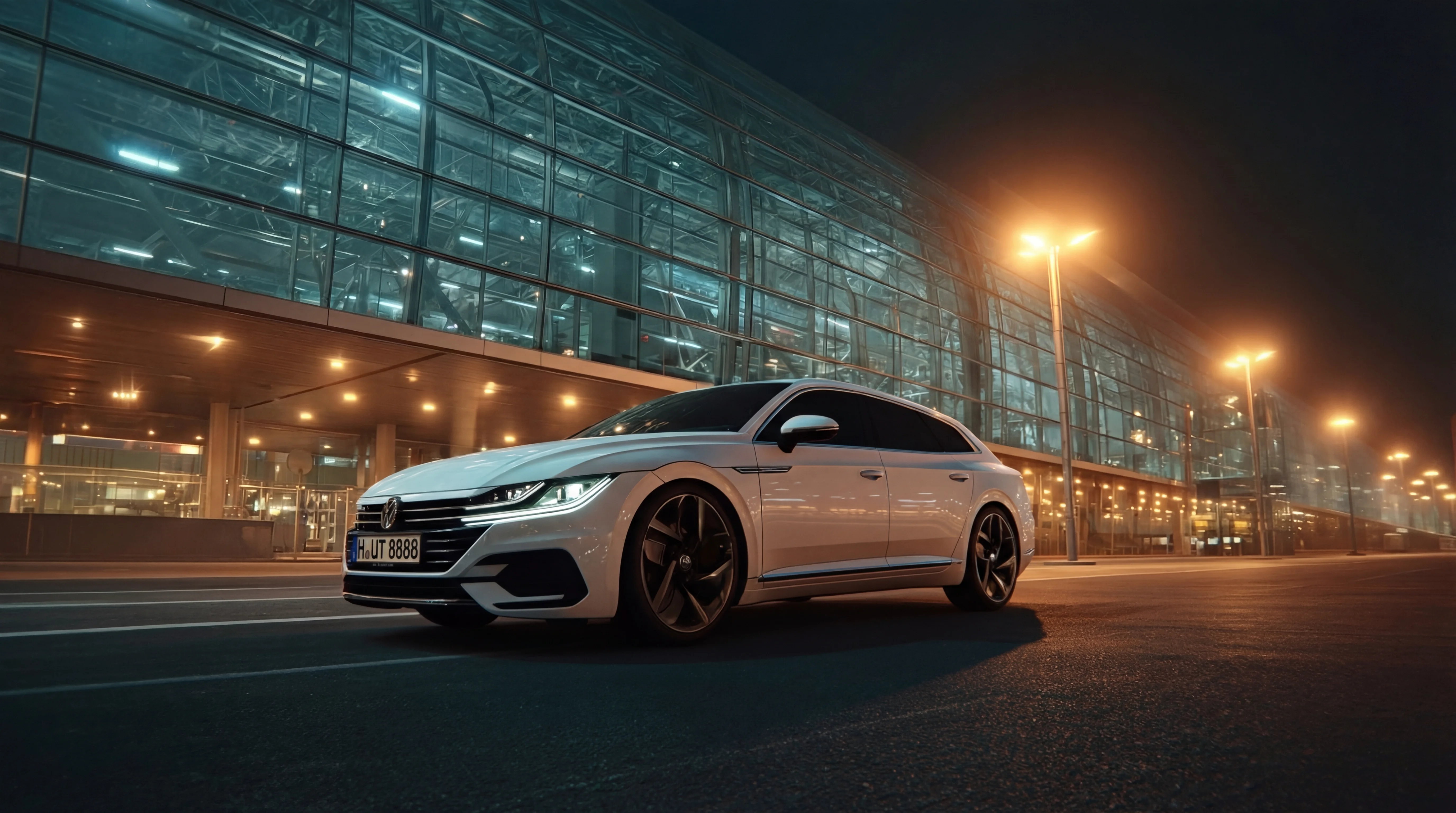 Ein weißer Volkswagen Pkw fährt nachts mit eingeschalteten Scheinwerfern auf einer Straße in der Stadt; im Hintergrund ist ein Flughafen zu sehen.