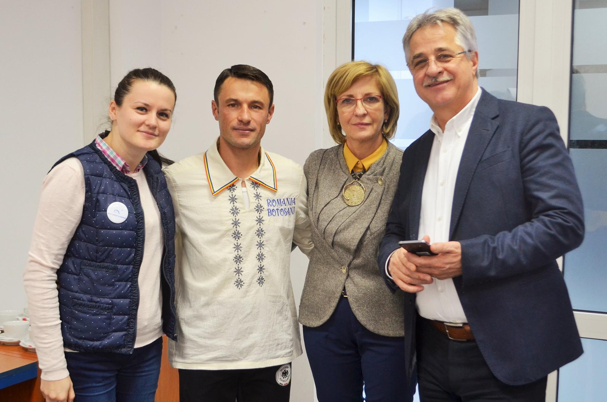 A young person with curly hair smiles at the camera while sitting in front of a laptop against a dark background.
