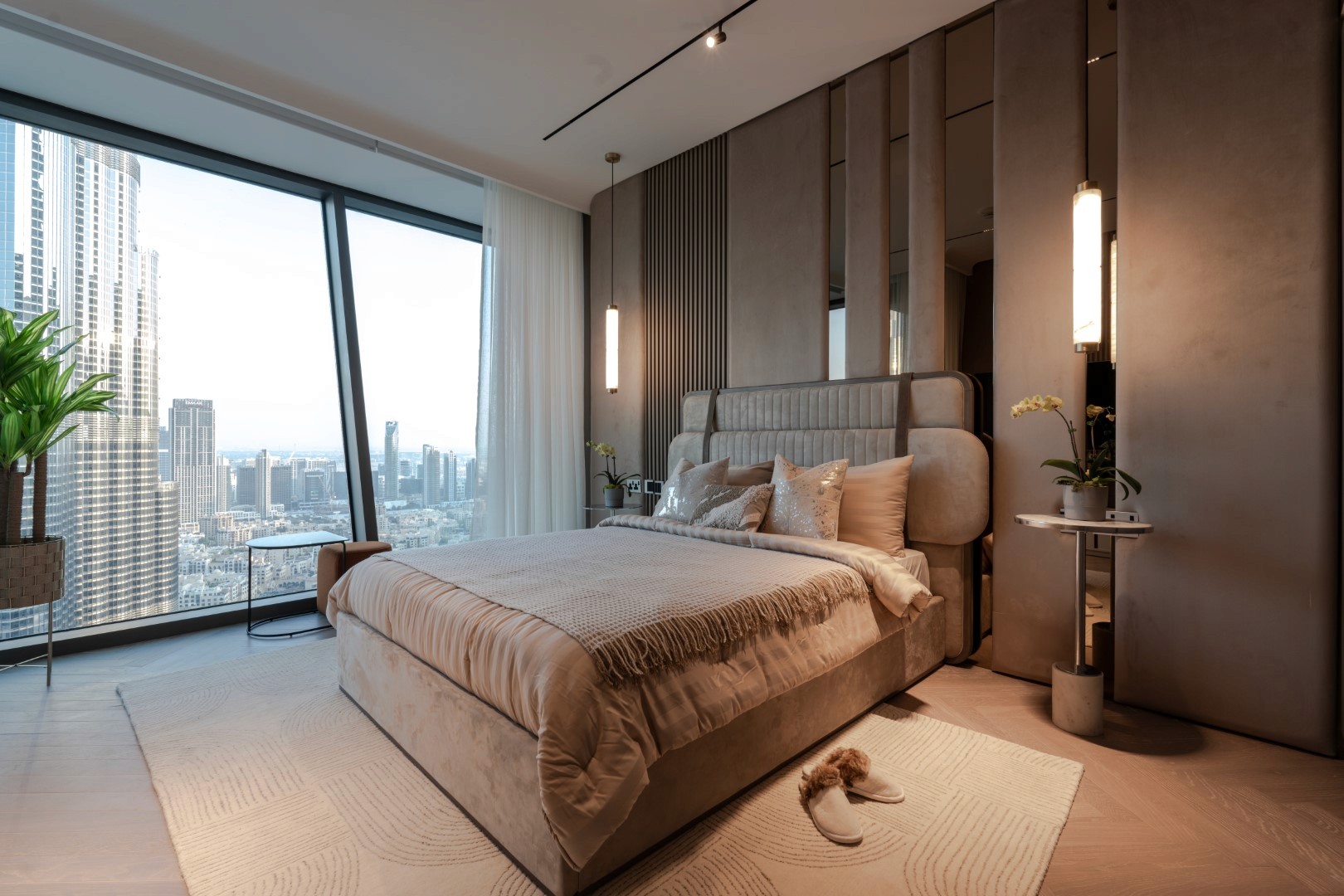 Luxurious modern bedroom in Burj Vista, Dubai with a Burj Khalifa view, featuring a plush beige bed with textured bedding, sleek floor-to-ceiling windows offering a stunning city skyline view, sophisticated vertical wall panels, ambient pendant lighting, and a green potted plant for a touch of nature.