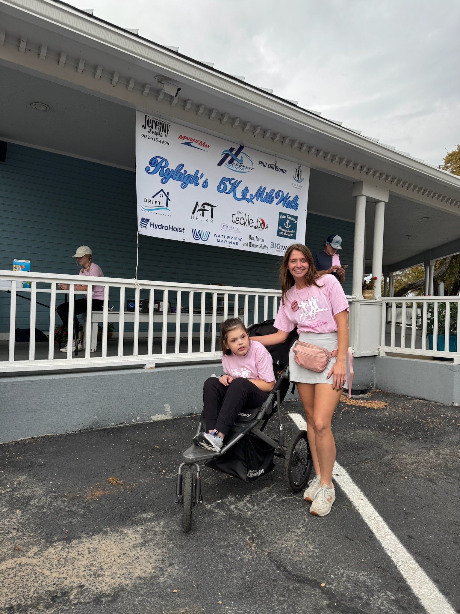 A woman wearing a pink t-shirt stands smiling next to a young girl in a stroller in front of a building with a large sign for a 5K and 10K race event, showcasing a sense of community participation and support for the event.