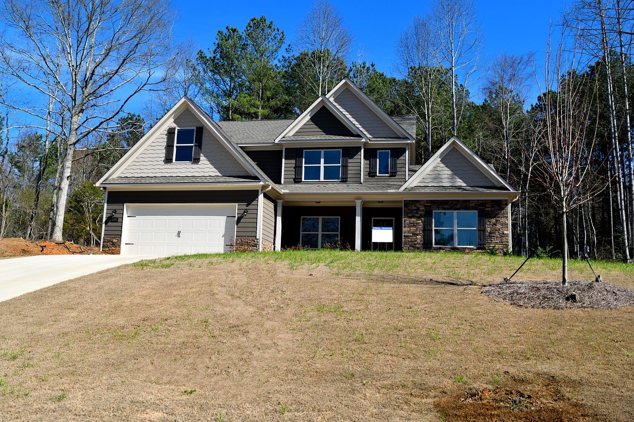 A large suburban house with a two-car garage, surrounded by bare trees and a slightly sloped, brown grassy lawn under a clear blue sky.