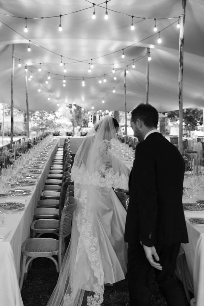 Sposi camminano lungo tavolo allestito con luci sospese, reportage di matrimonio in villa con atmosfera elegante e intima