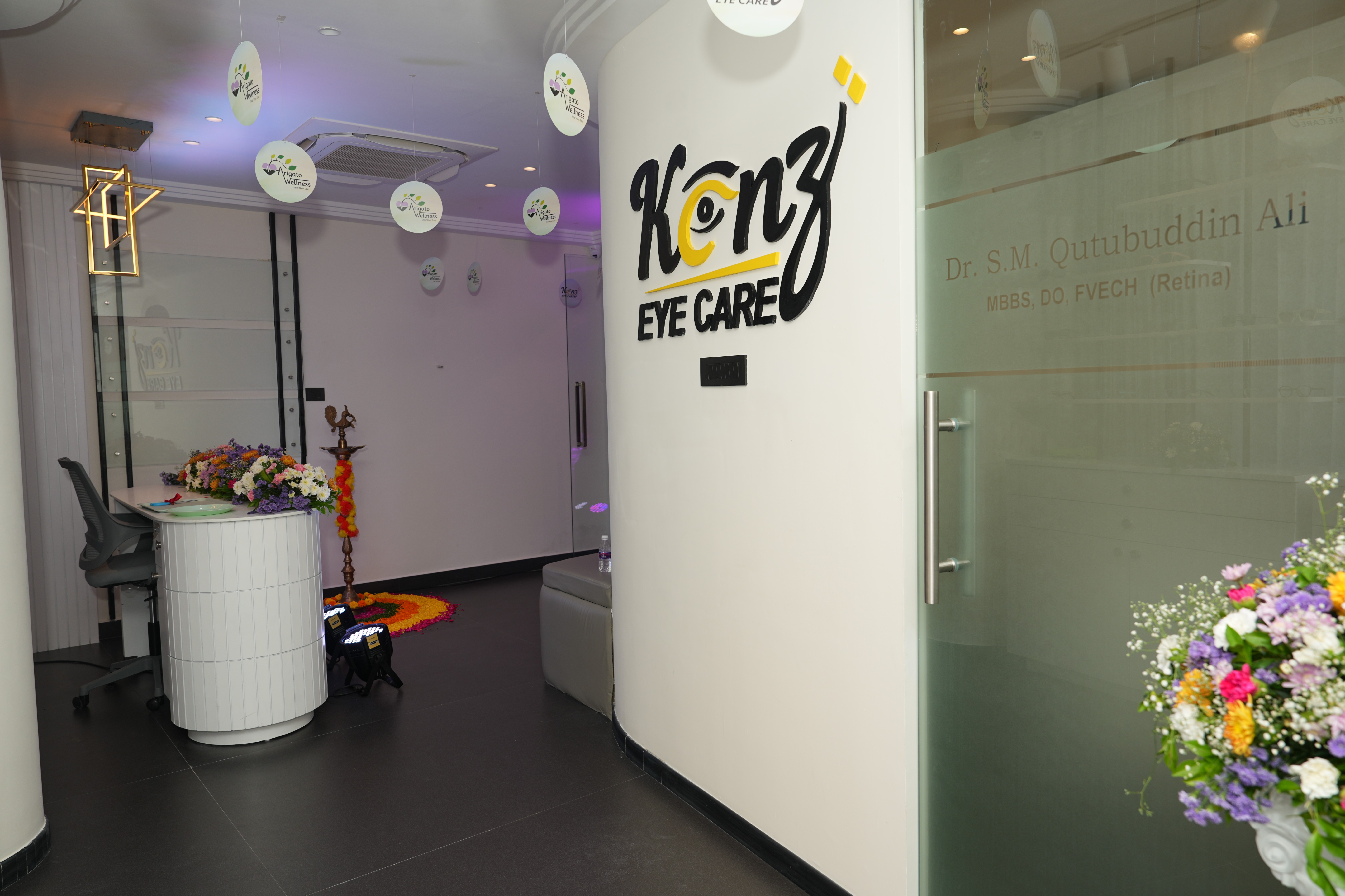 The image shows the reception area of a modern eye care clinic named "Kenz Eye Care" located in Kokapet, Hyderabad. The clinic features a welcoming and clean interior with white walls and a large, stylized black and yellow logo. On the right, a frosted glass door leads to a consultation room, with the text "Dr. S.M. Qutubuddin Ali MBBS, DO, FVEH (Retina)" visible. The reception desk is decorated with fresh flowers, and traditional Indian lamps are placed on the floor, suggesting an inauguration or a festive occasion. The overall impression is professional and inviting, appealing to potential clients looking for an eye check-up, retina specialist, or general eye care services in and around Kokapet, Hyderabad