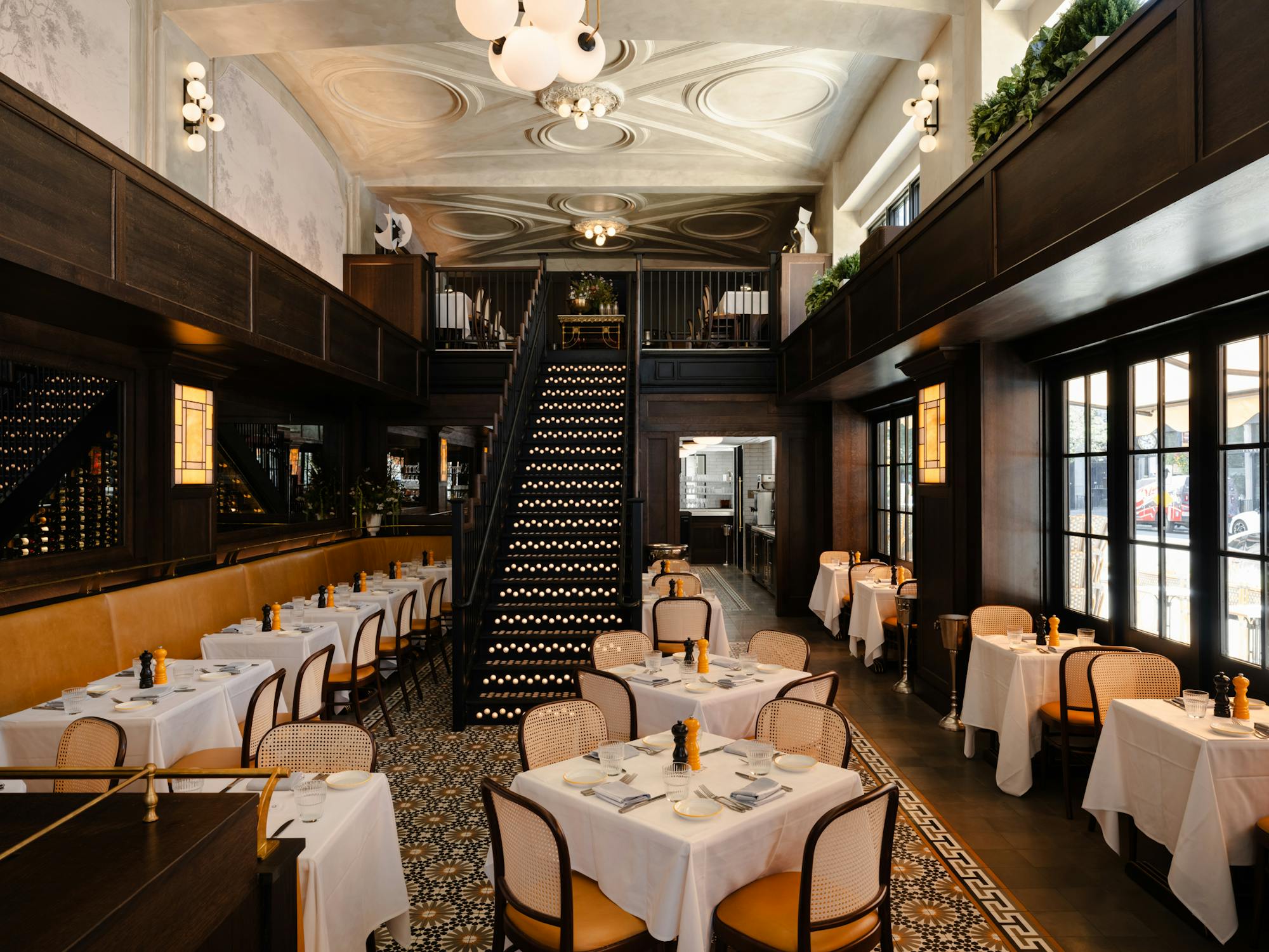 Dining room with white-clothed tables, cane chairs, banquettes and a staircase at Chouette