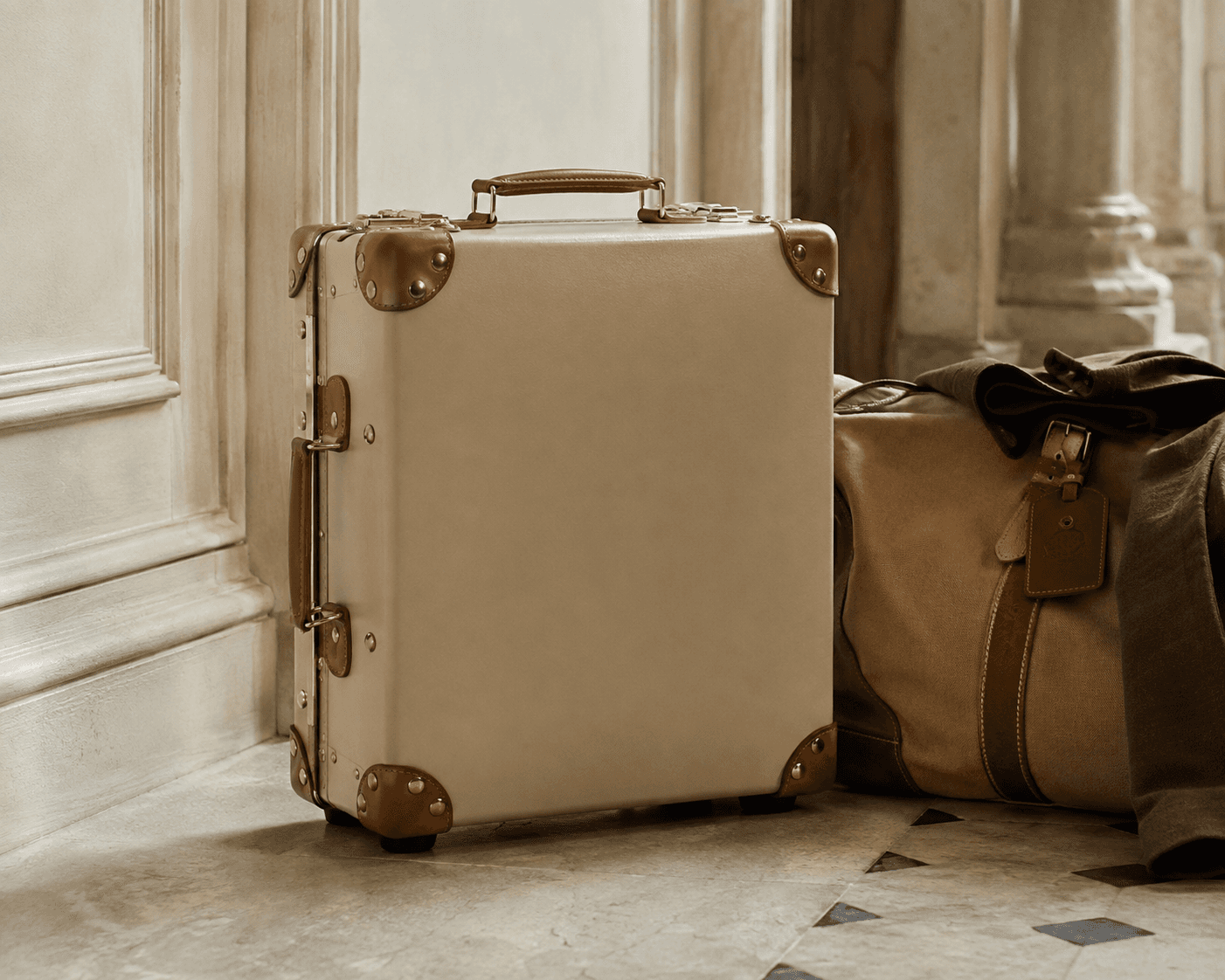 A vintage-inspired luxury travel scene featuring a beige rigid suitcase trunk with tan leather corner protectors, matching leather handles, and brass latches standing upright on a stone tile floor. Beside it sits a tan canvas weekender duffel bag with leather trim and a brown jacket draped over the top against a classic architectural interior backdrop. It depicts a Globe-trotter suitcase in a pannelled cream room.