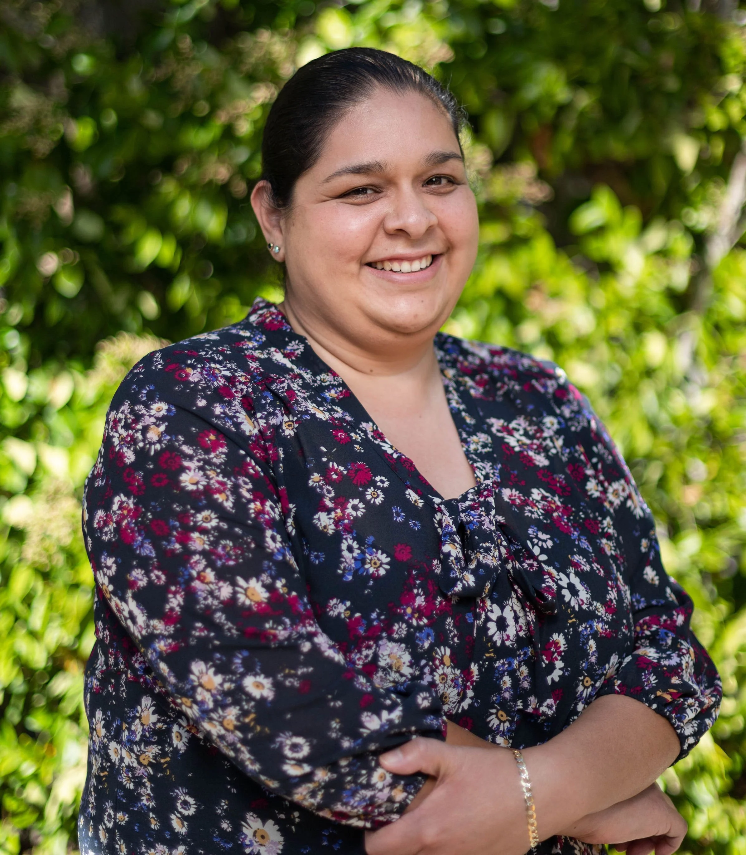 Uma pessoa sorri calorosamente para a câmera, em pé ao ar livre diante de um cenário exuberante de folhagem verde vibrante, vestindo uma blusa com estampa floral e os braços cruzados.